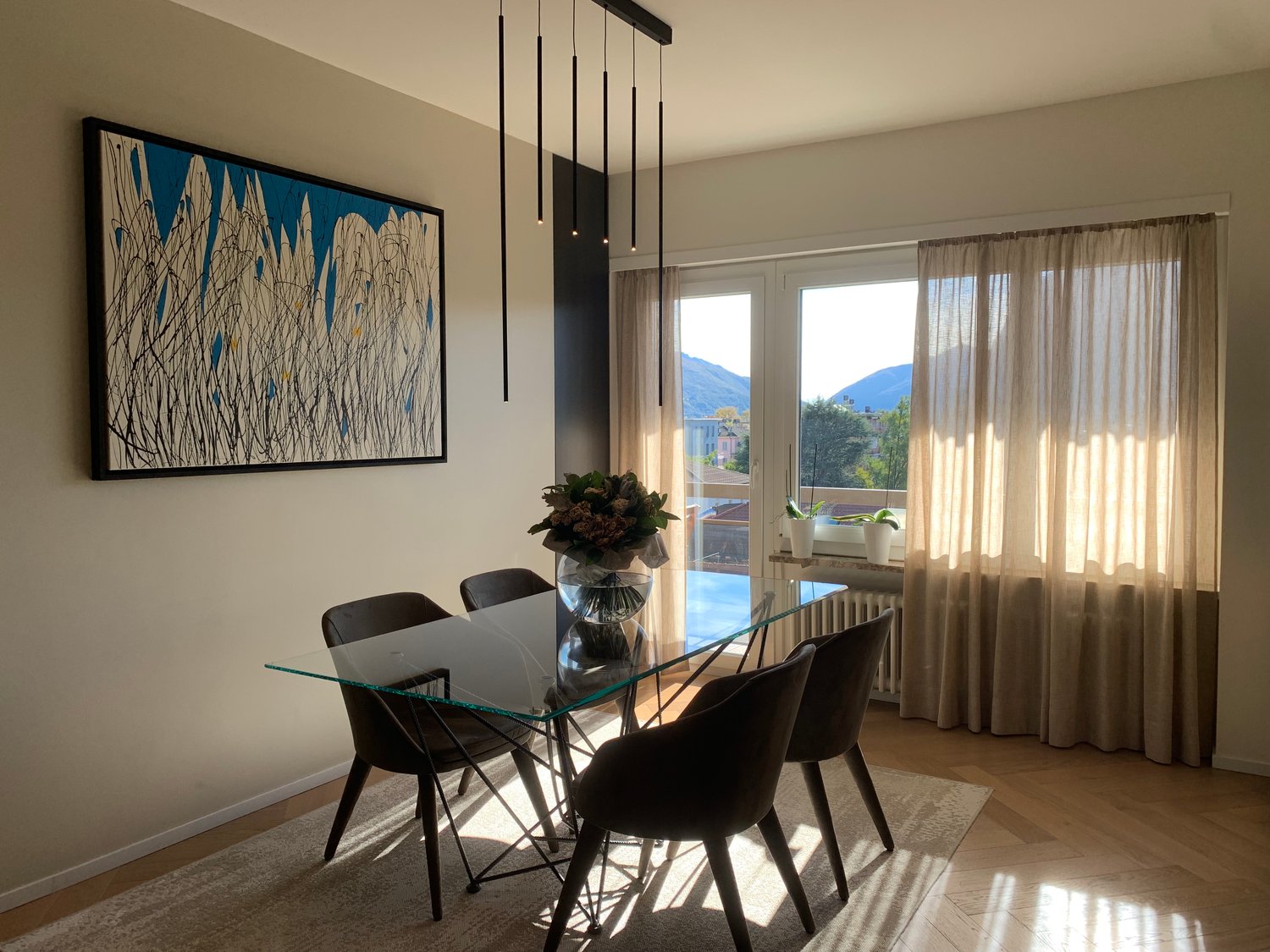 Spacious dining area with a glass-top dining table, black chairs, and a large abstract artwork on the wall. The room features large windows with a scenic view of the mountains outside.