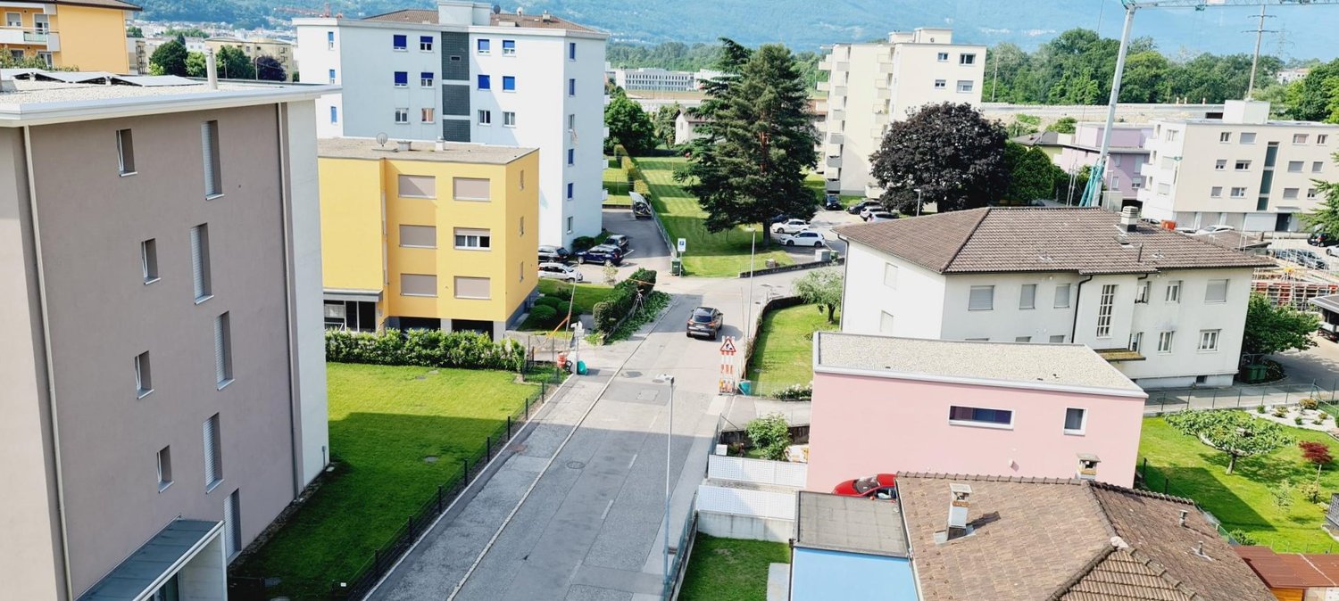 multi-story buildings, paved road, parked cars, green grass, trees, mountains in the distance