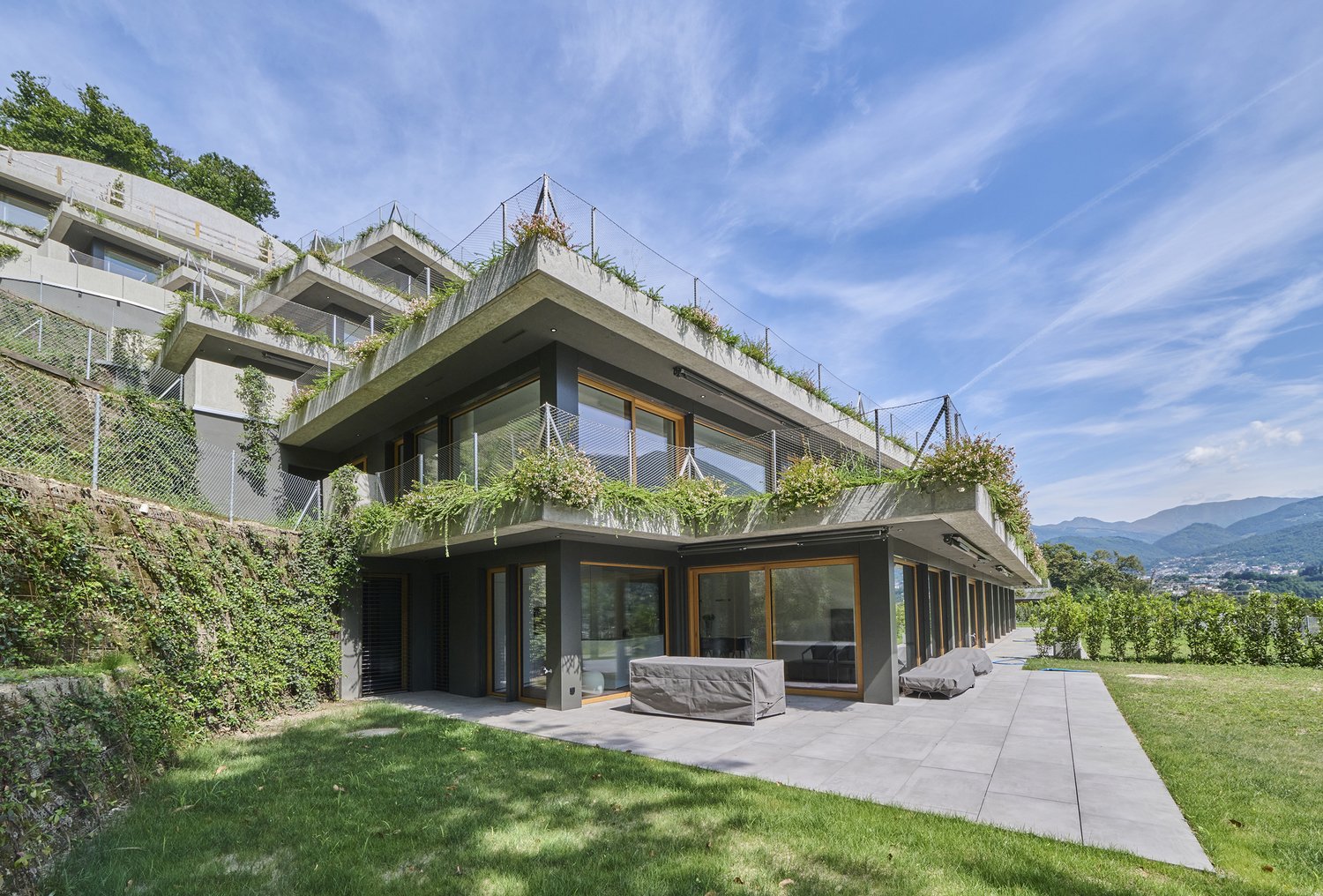 Modern apartment house with large glass windows and terraces, multiple levels, roof garden, and concrete structure.