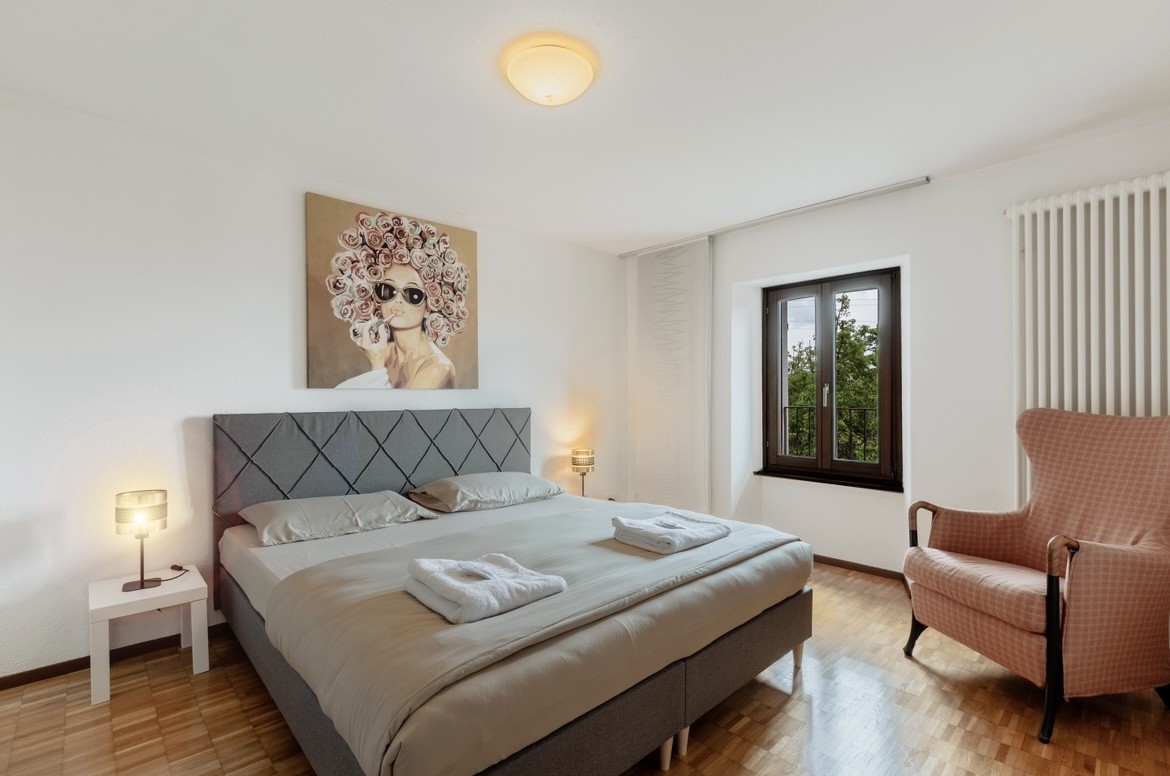 Spacious bedroom with a large bed, gray upholstered headboard, artwork on the wall, and a window overlooking the outdoors.