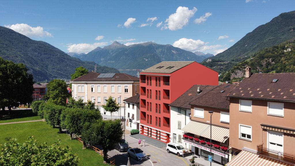 Multiple buildings, large mountains in the background, large park with trees, cars parked on the road.