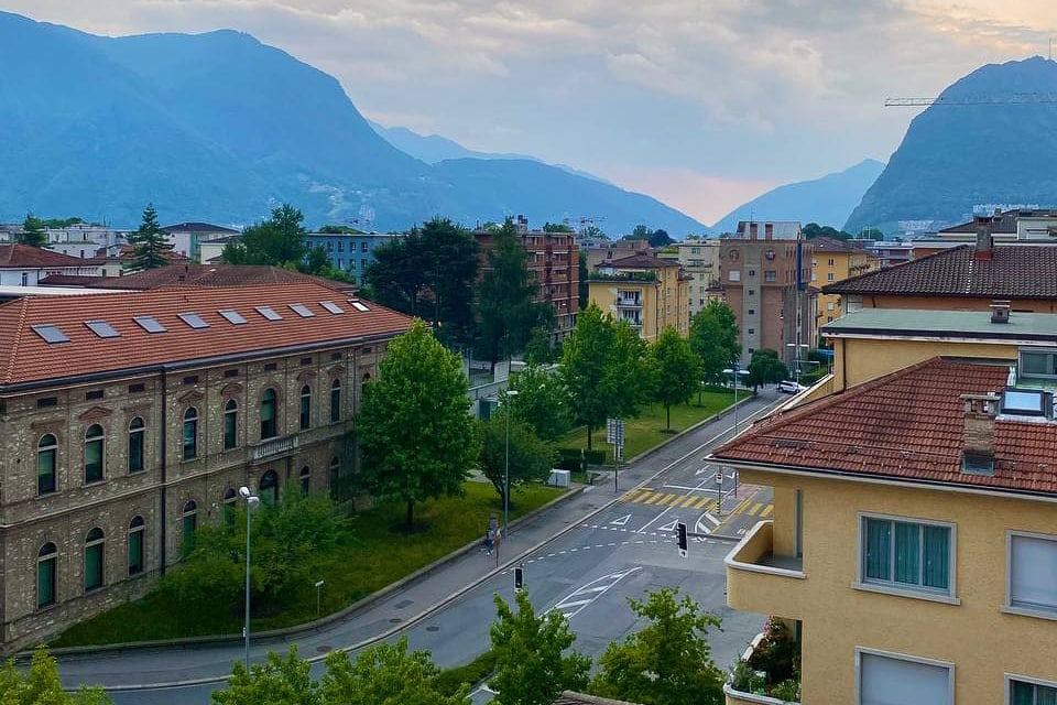 Aerial view of an urban area with multiple buildings, green trees, and mountains in the background.