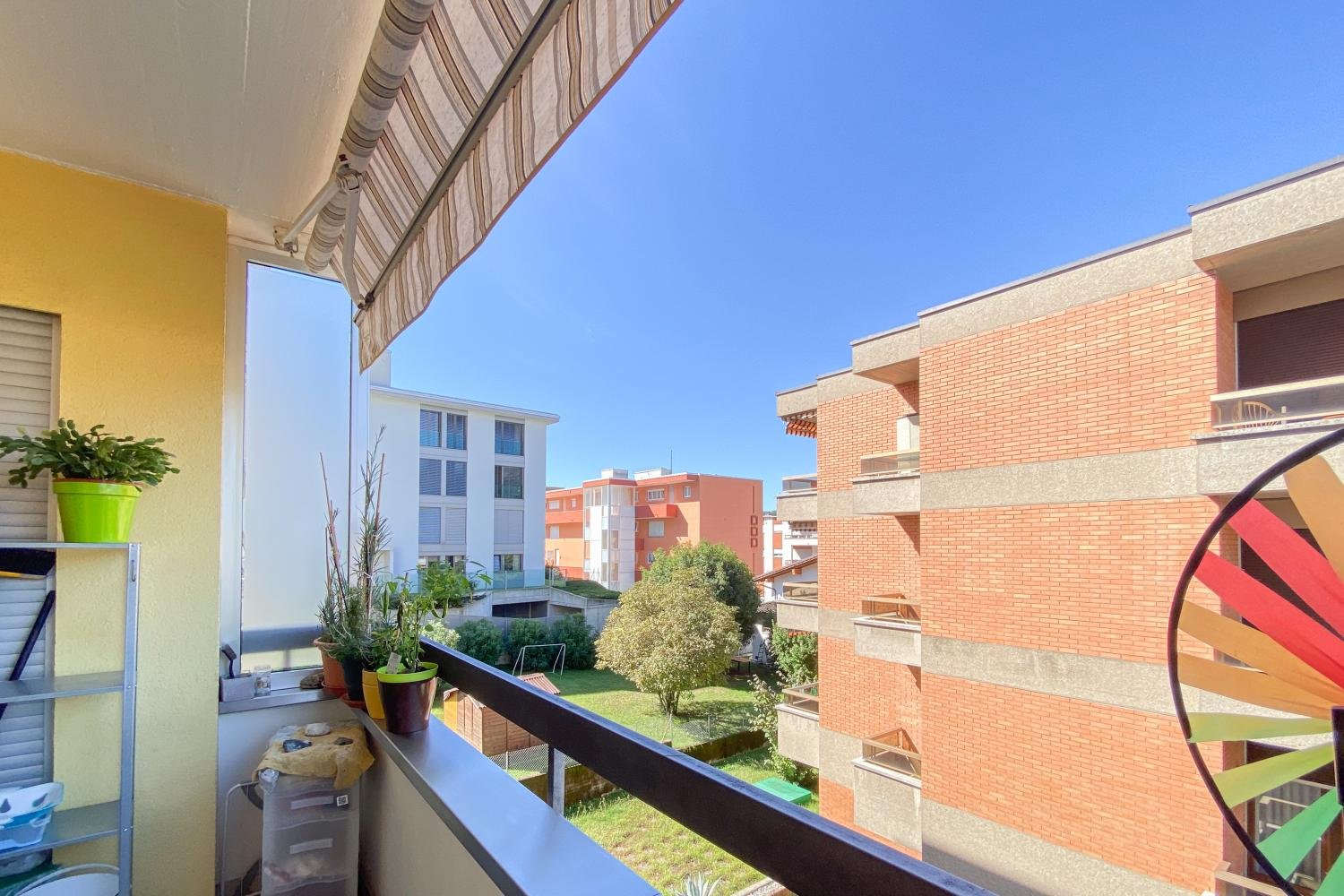 balcony, railing, plant pots, wind spinner, distant buildings, greenery, blue sky