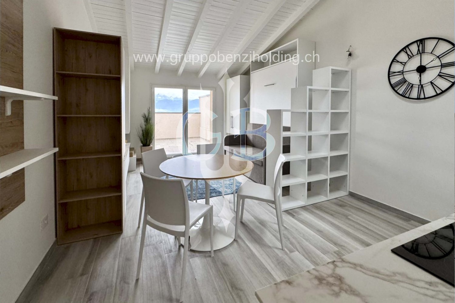 A room with white and gray furniture, including a desk and chairs, a round table, shelves, and a clock on the wall. There is a glass door leading to a balcony with a view of the outdoors.