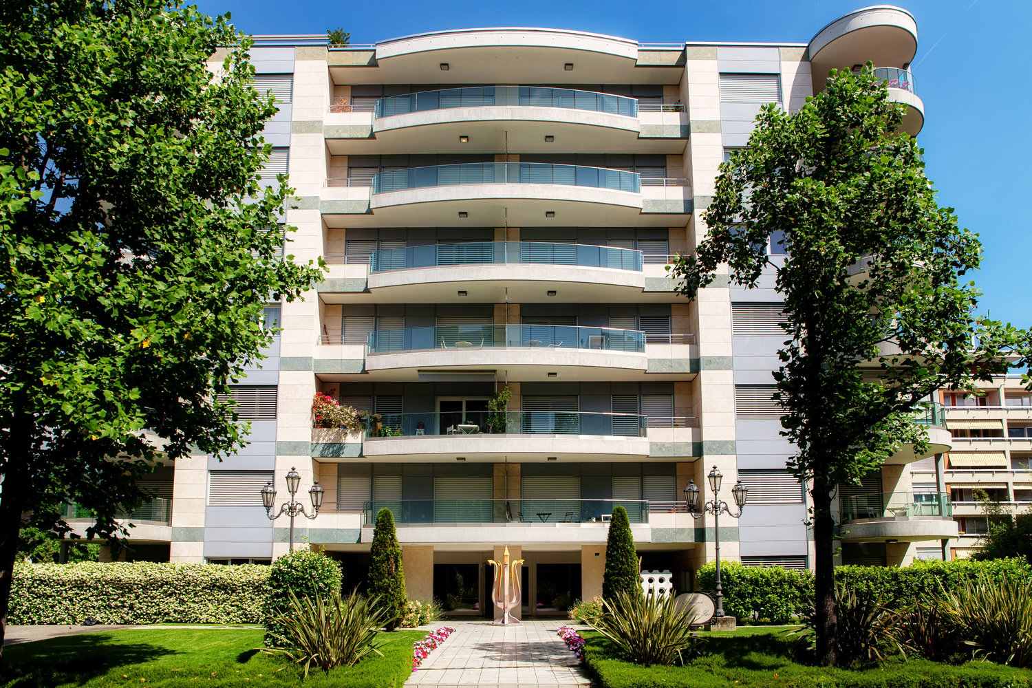 multi-story building, balconies on every floor, entrance with arch, multiple plants and trees