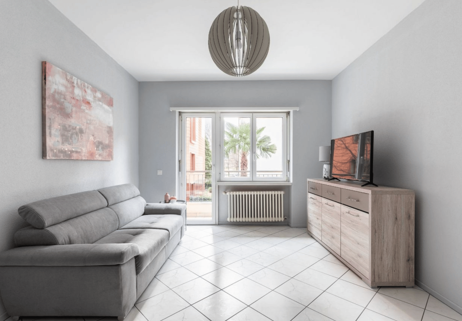 modern living room with gray couch, white tile floor, large windows, balcony, TV, wooden cabinet, radiator