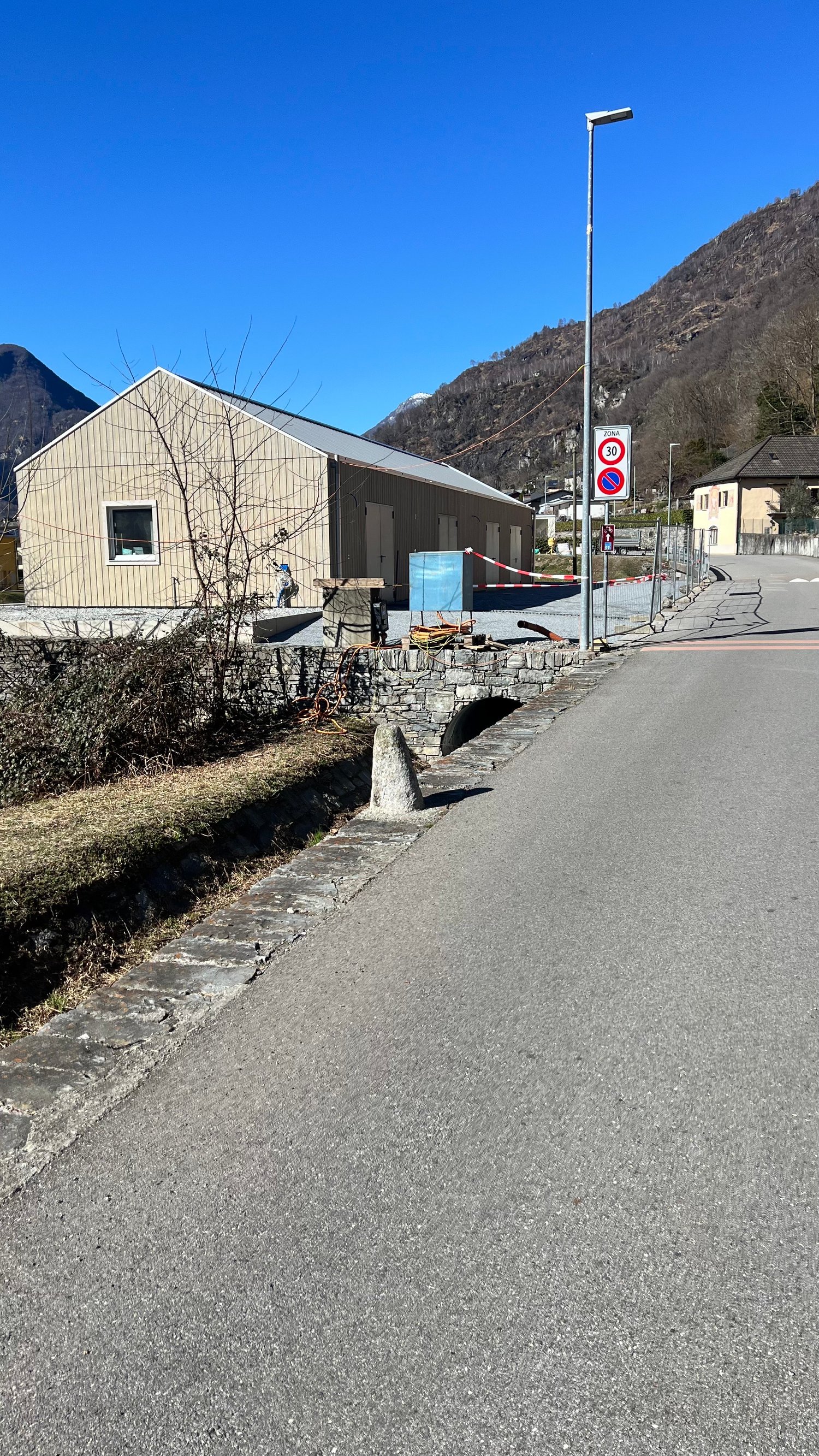 A single-story gray building with a slanted roof, surrounded by a stone wall and located in a mountainous area. There is a street with a speed limit sign visible in the foreground.