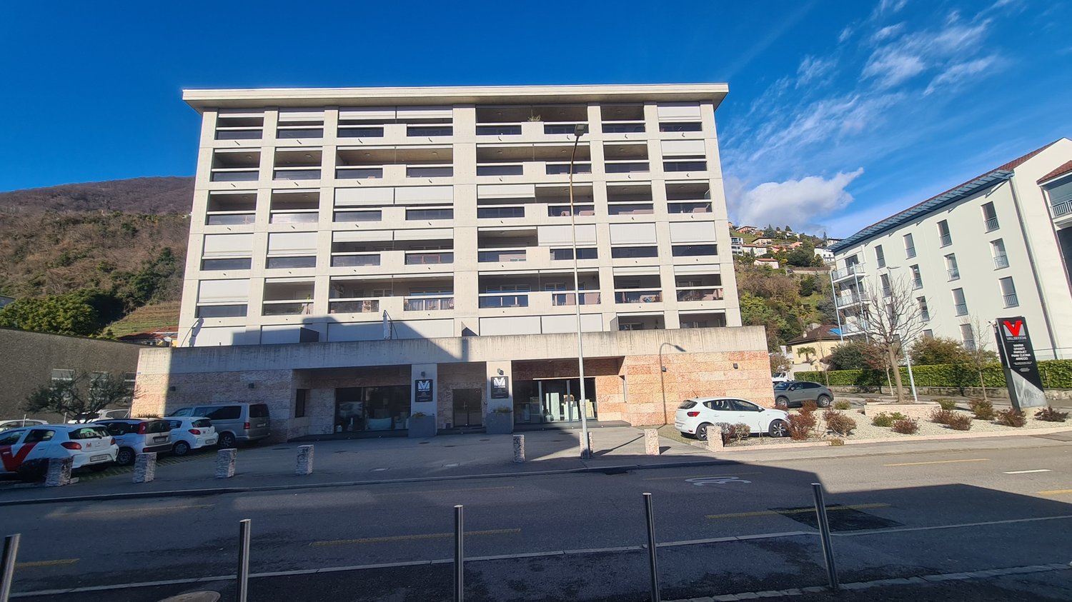 High-rise building, concrete, multiple windows, cars parked, streetlight, front entrance, small bushes, bollards, flat road.