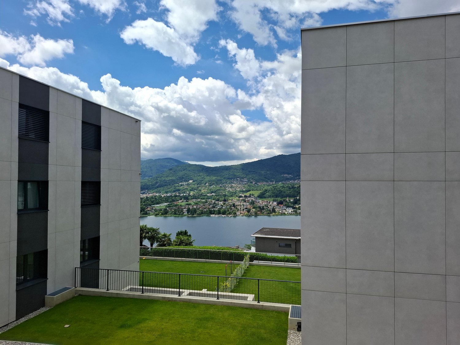 Exterior view of two-story building, large windows, fenced roof garden with lush grass, lake and town in the background
