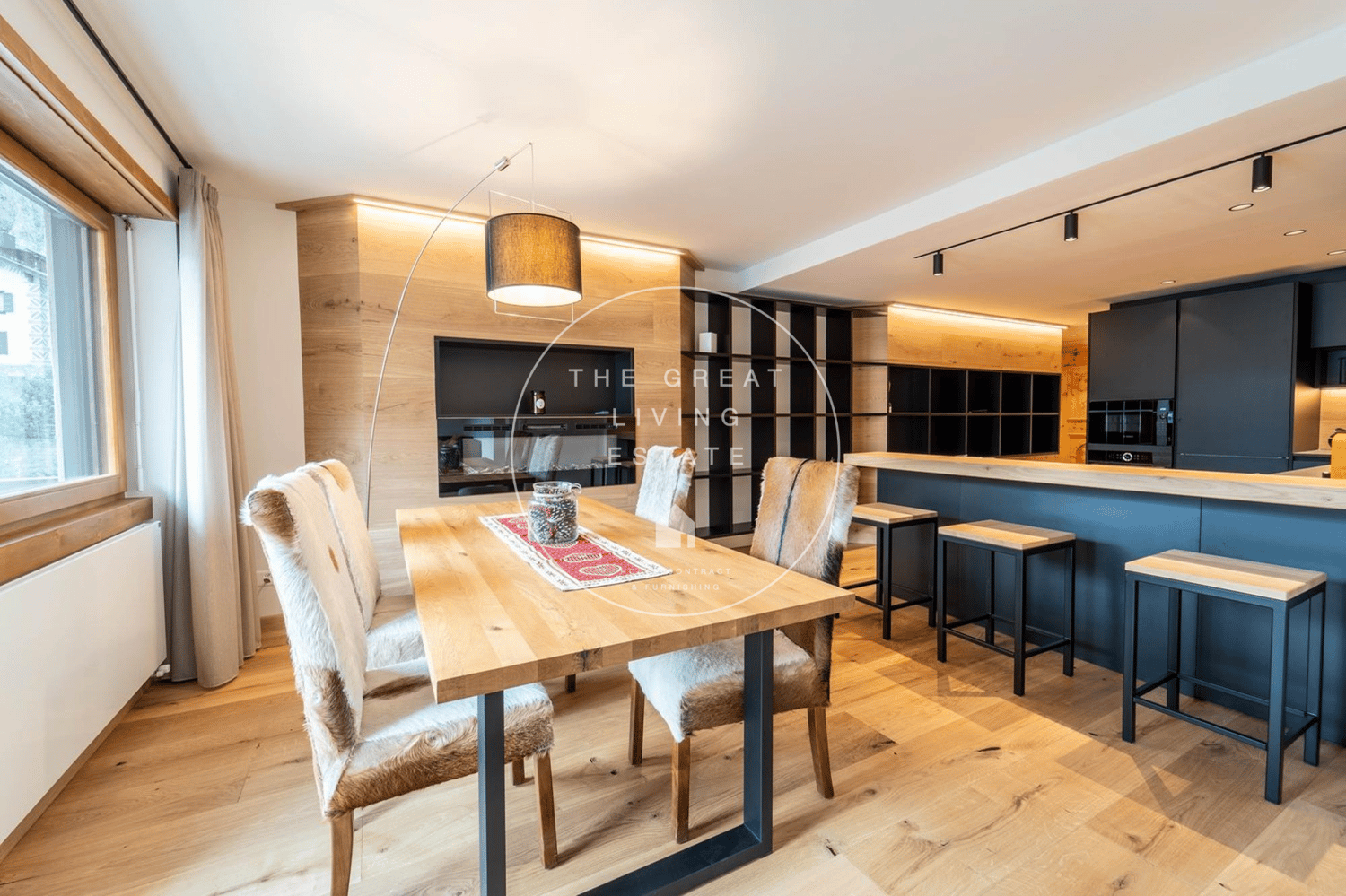 The image shows a modern, open-plan living room and kitchen area. The space features a large wooden dining table with fur-covered chairs, a black kitchen with wooden accents, and a fireplace on the wall. The room has a cozy and inviting atmosphere with wa