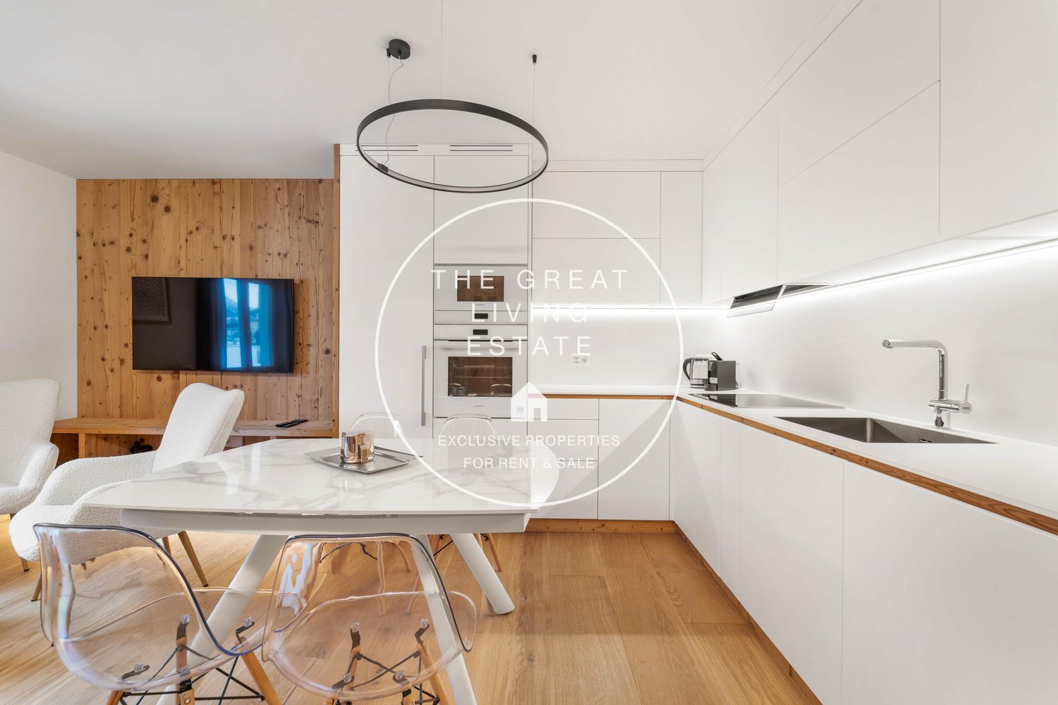 The image shows a modern and minimalist apartment interior. The living room has a TV mounted on a wooden wall panel, and a circular pendant light above a dining table with transparent chairs. The kitchen area has a sink and countertop with wooden accents.