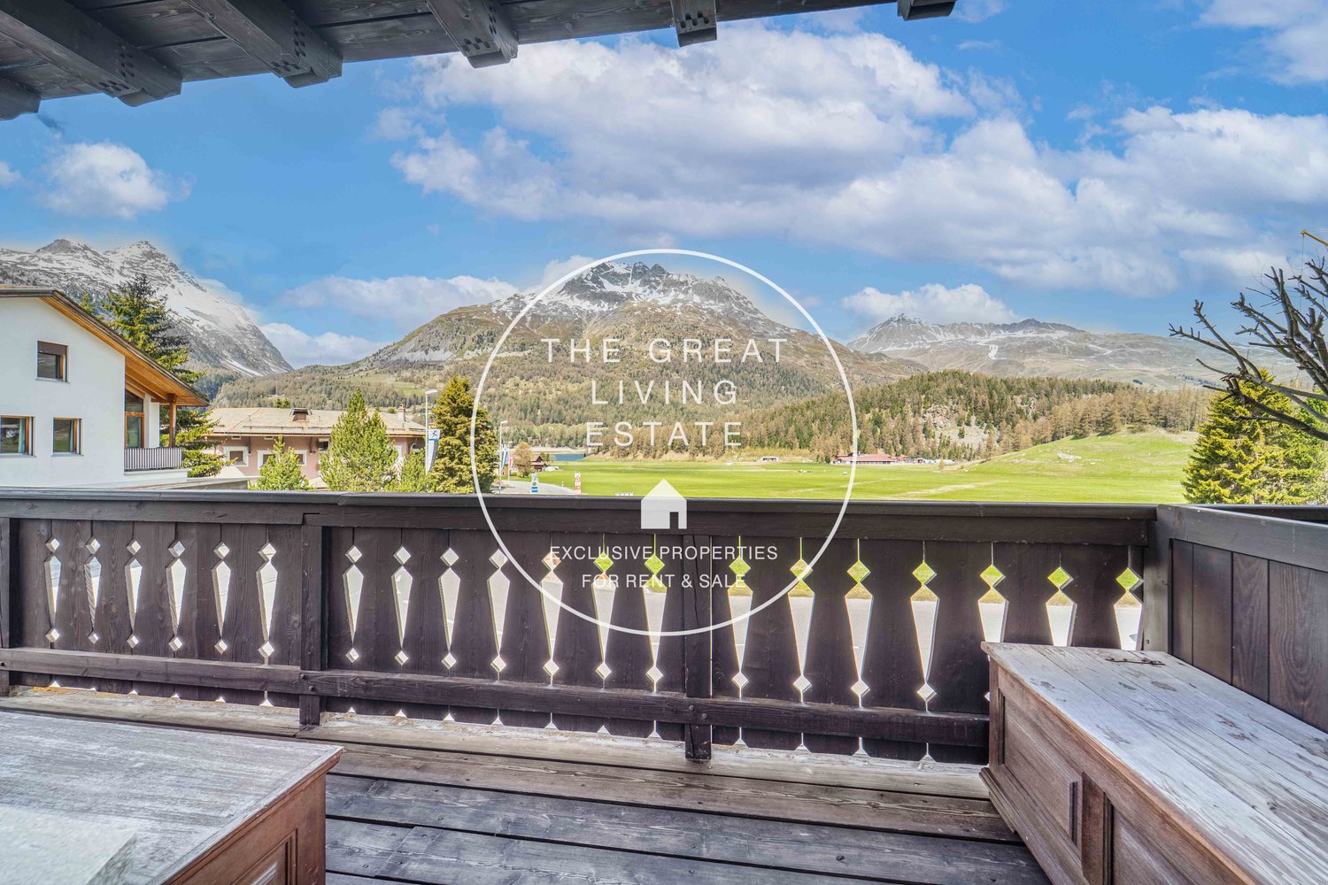 The image shows a scenic mountain landscape with a wooden balcony or terrace in the foreground. The balcony has a railing with a decorative pattern and there is a sign that says 'The Great Living Estate' with the text 'Exclusive Properties for Rent & Sale