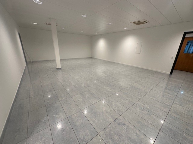 A room with white walls, ceiling, and tiled floor, and lights installed in the ceiling. There is a pillar in the middle of the room and an exit door at the right end.
