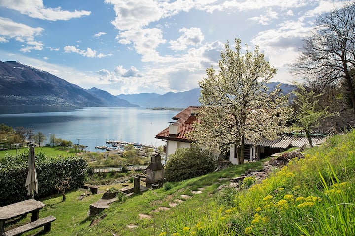 Two-story house with a red tile roof, surrounded by a lush garden with blooming trees and flowers. The house overlooks a scenic lake with mountains in the background. There appears to be a balcony or terrace on the upper level of the house, offering a bea