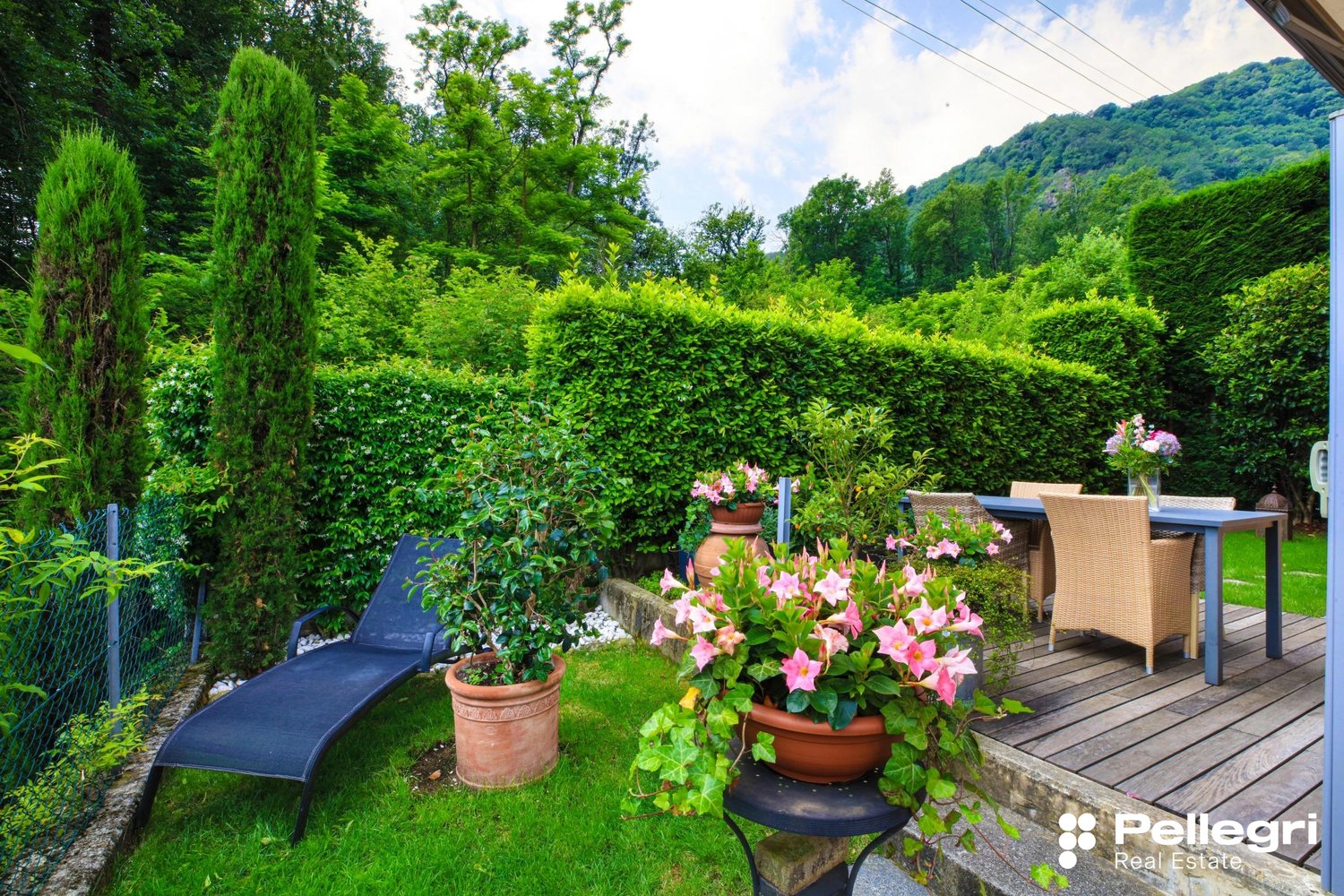 Lush green garden with tall trees, hedges, and potted plants. Wooden deck with outdoor furniture and a fireplace. Scenic mountain view in the background.