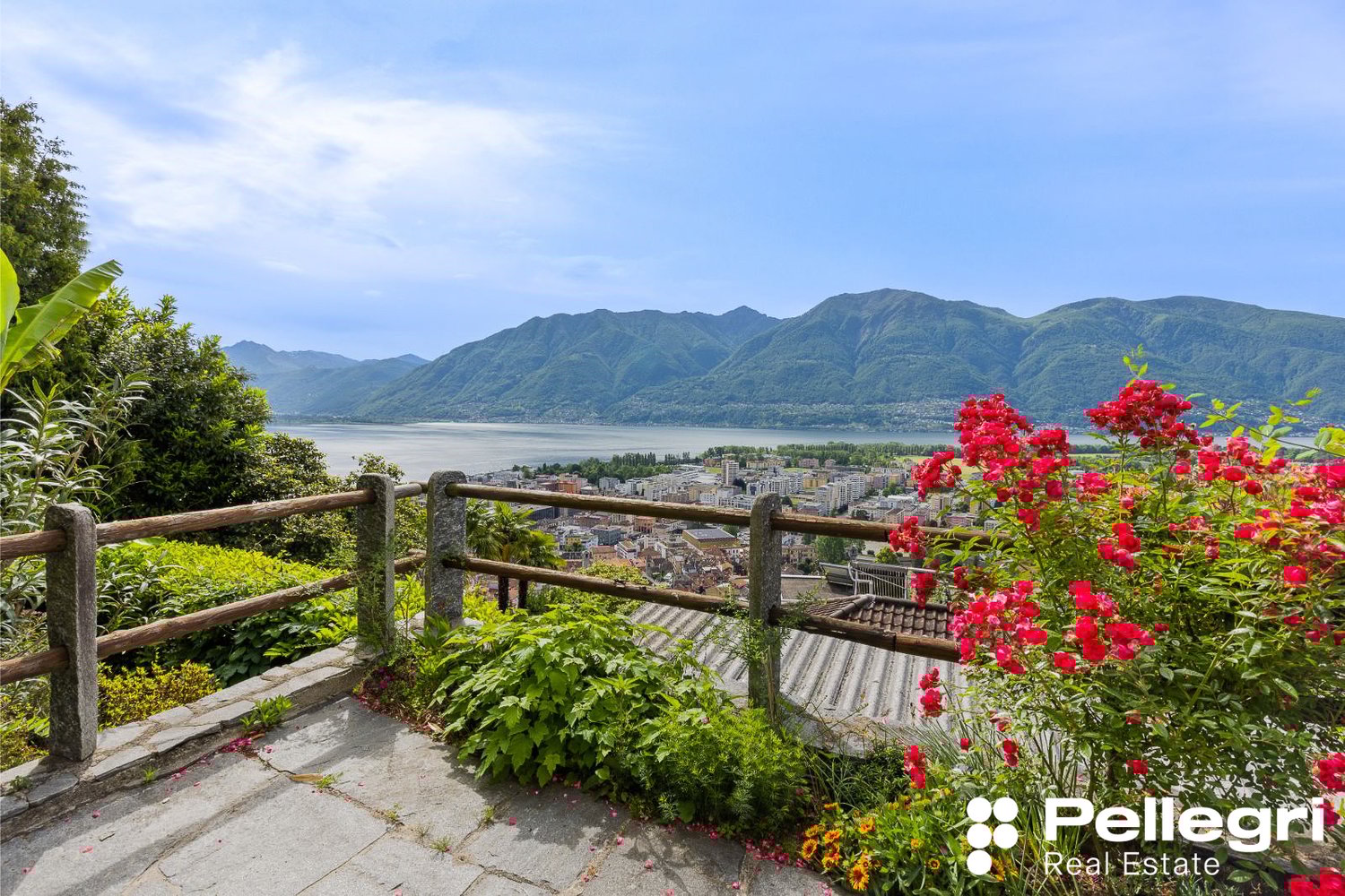 Scenic view of a lakeside town surrounded by mountains, with a wooden railing and lush greenery in the foreground featuring vibrant red flowers.