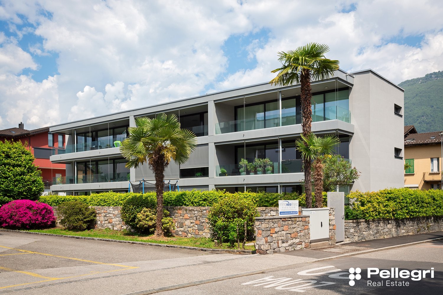 Modern multi-story apartment building with glass facades, balconies, and palm trees in the landscaping. The building has a stone wall at the front with the Pellegri Real Estate logo.