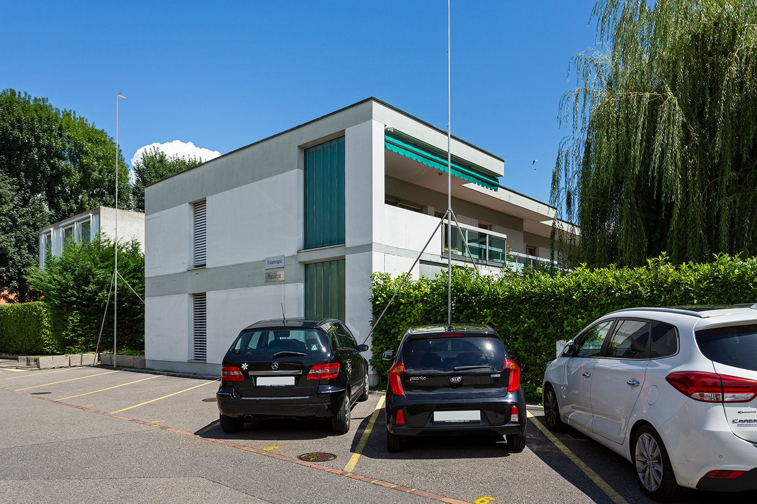 2-story commercial building with a modern, minimalist design. The building has a gray exterior with green accents and large windows. There is a parking lot in front of the building with several cars parked.