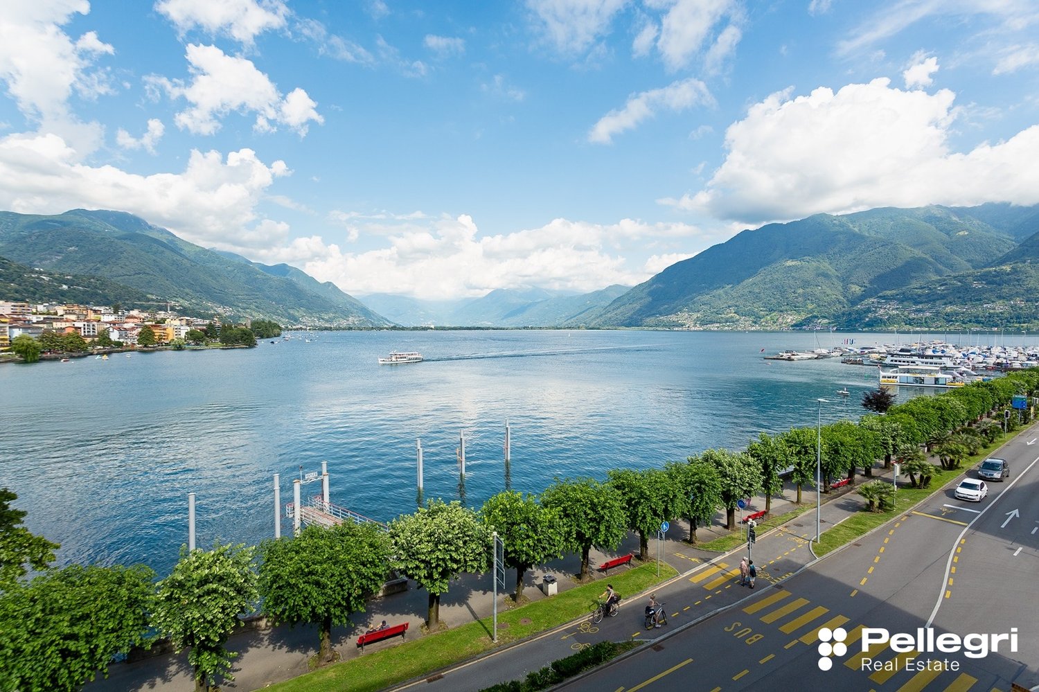 The image shows a scenic view of a lake surrounded by mountains. The lake is dotted with boats and yachts, and the shoreline is lined with trees and buildings. The sky is blue with fluffy white clouds, creating a picturesque and serene atmosphere.