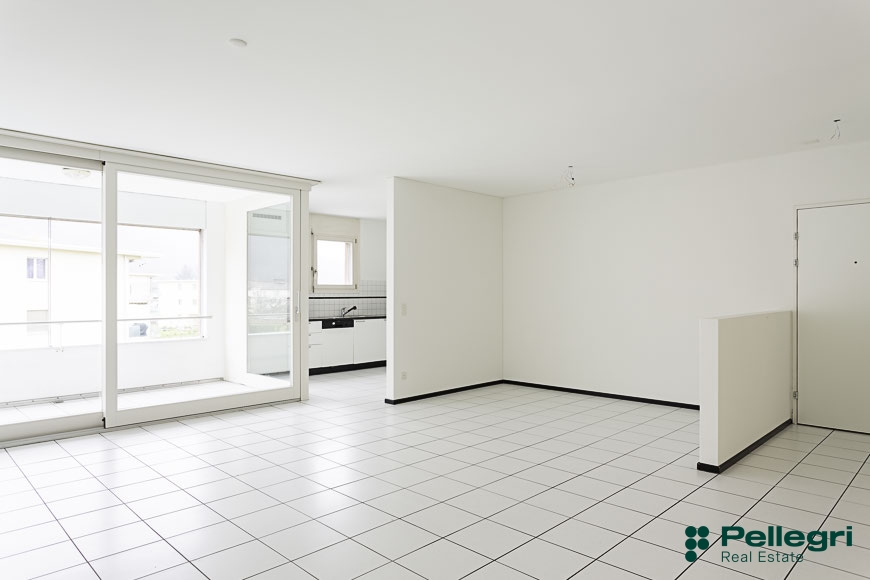 Open floor plan with large windows, tiled flooring, and a kitchen visible in the background.