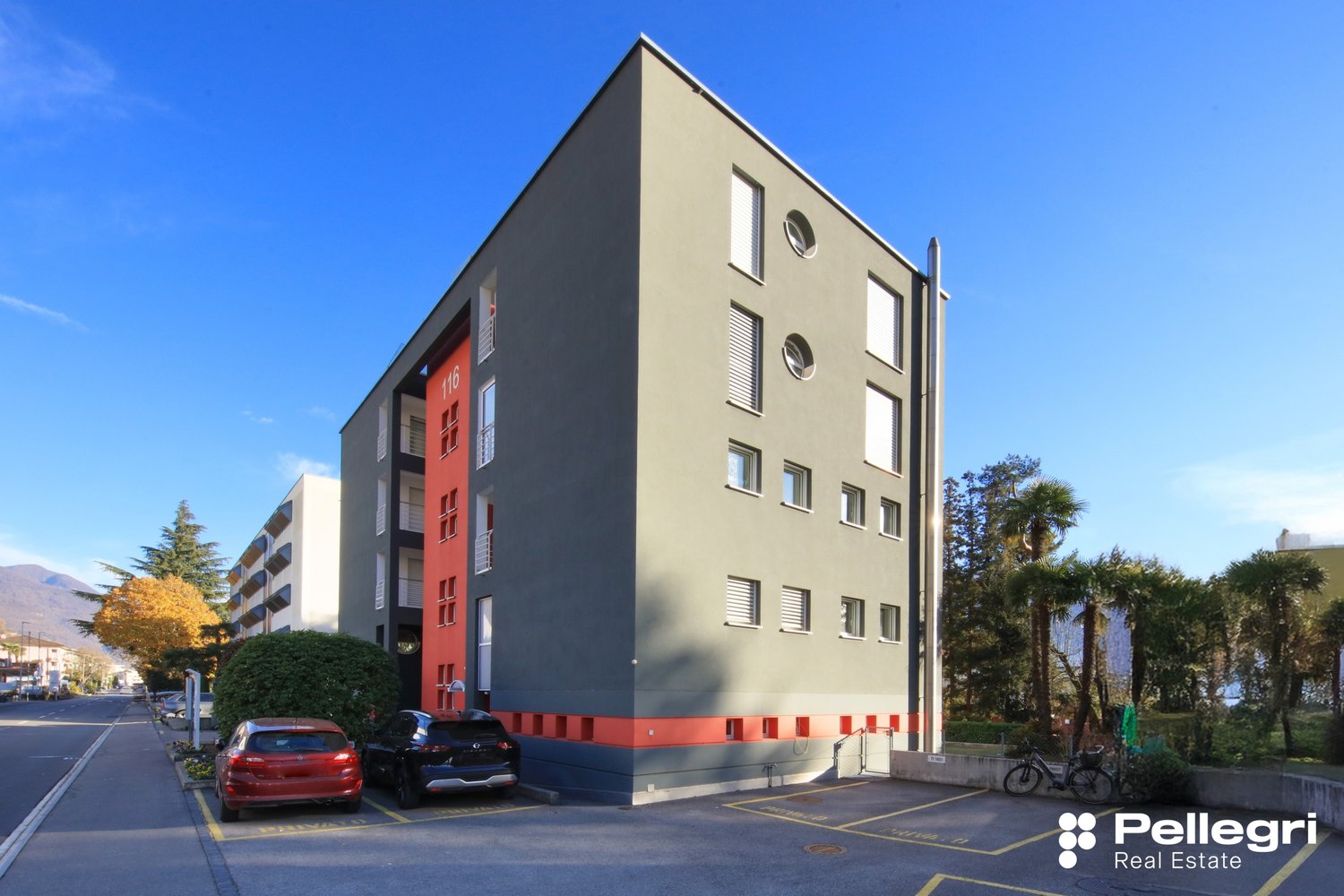 Gray and red building, two cars parked, trees and greenery around, parking lot in front