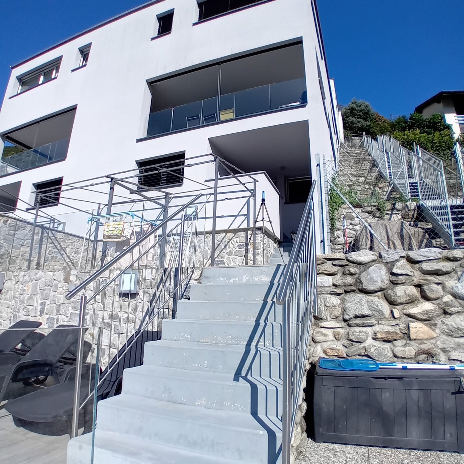 Modern white building, multiple balconies, staircases leading up and down, large windows, two lounge chairs on ground floor