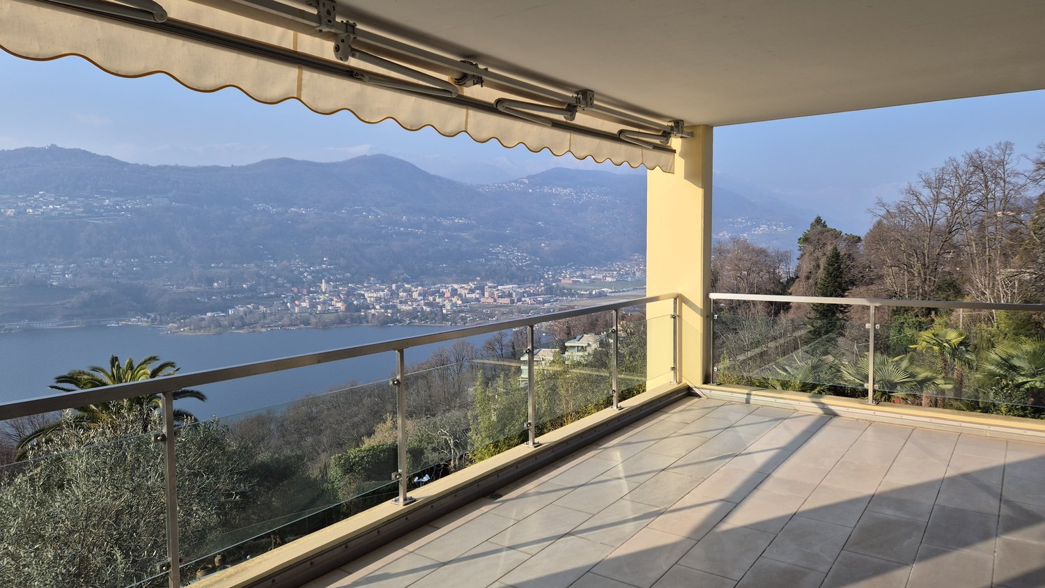 Spacious balcony with a retractable awning, offering a panoramic view of the surrounding mountains and lake. The balcony has a tiled floor and glass railing, providing an unobstructed vista of the scenic landscape.