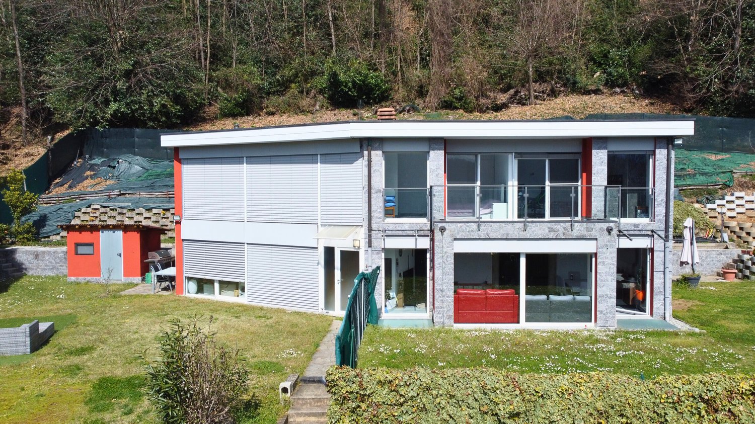 Two-story modern house with a flat roof, large windows, and a garage. The exterior is made of a combination of white siding and stone. There is a small paved area in front of the house that could be used for parking. The house is surrounded by a grassy ya