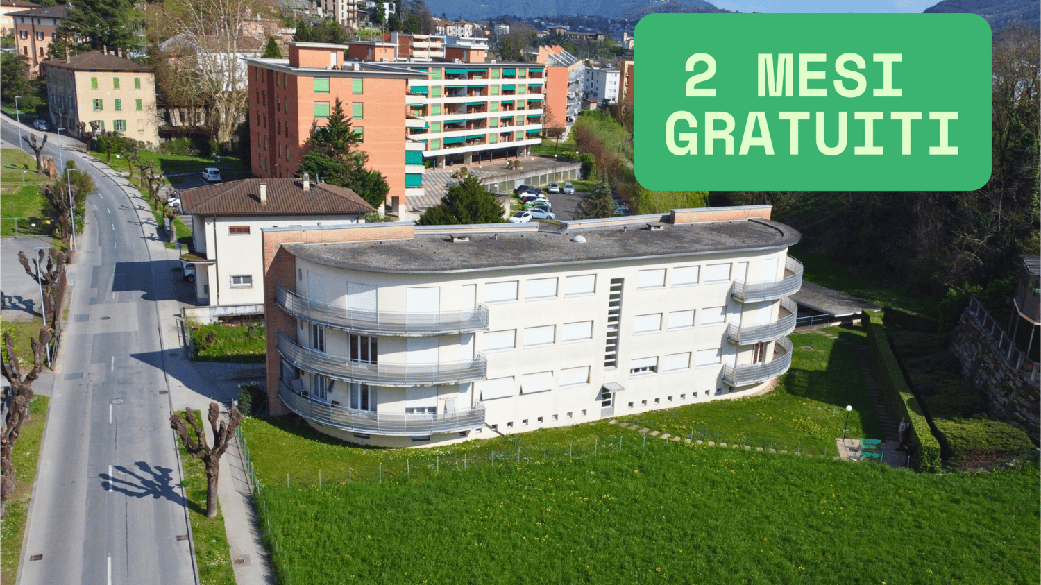 Multi-story apartment building with curved, rounded architecture, balconies, and green spaces surrounding it. The building appears to be located in an urban area with other buildings and roads visible in the background.