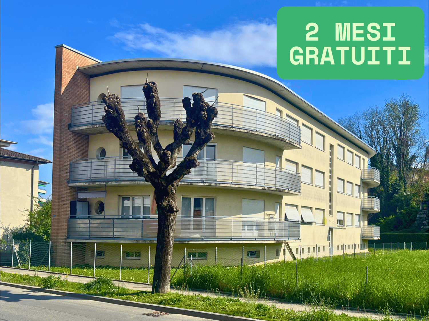 3-story apartment building with balconies, yellow exterior, surrounded by green grass and trees