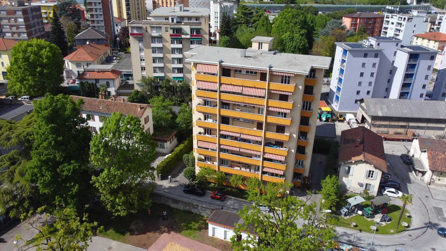 Multi-story apartment building with yellow and red balconies, surrounded by trees and greenery, with other residential buildings in the background.