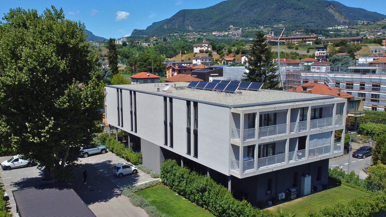 Multi-story apartment building with balconies, parking spaces, and solar panels on the roof. The building is surrounded by trees and mountains in a scenic, mountainous area.