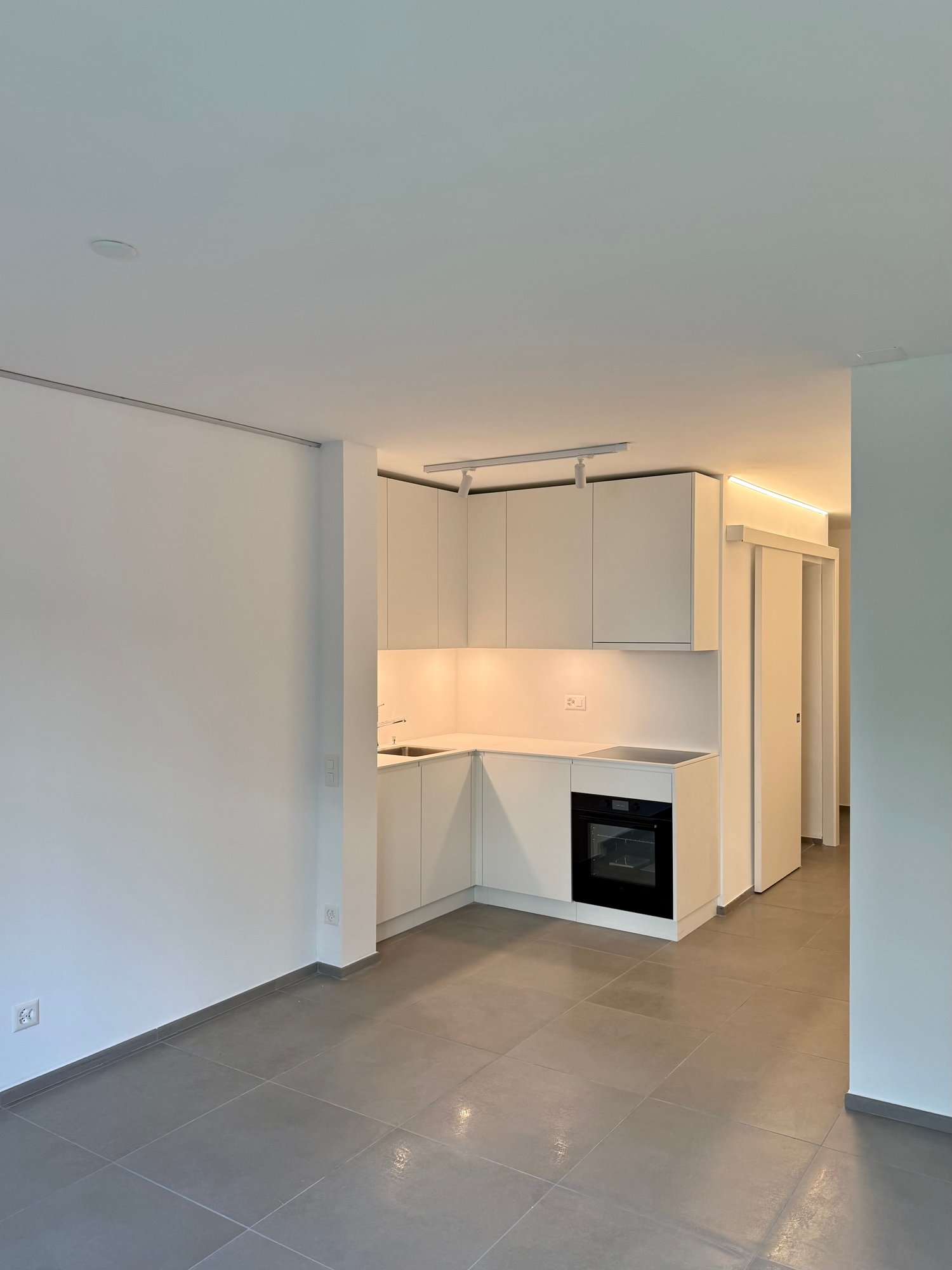 Modern kitchen, white cabinets, tiled flooring, integrated oven and stove, sink, ceiling track lighting, power outlets.