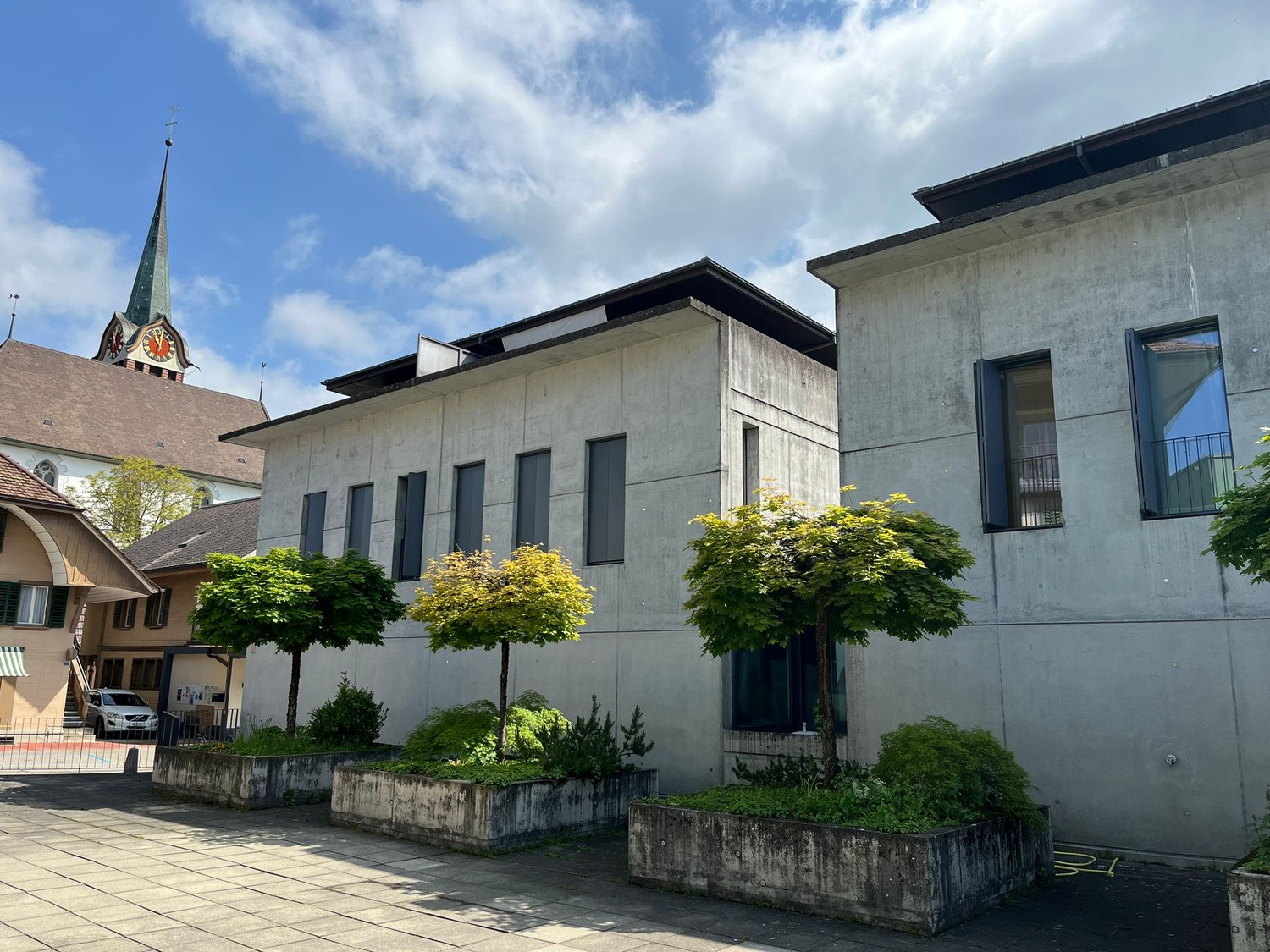 A large concrete building with a church spire in the background. The building has several windows and is surrounded by trees and landscaping.