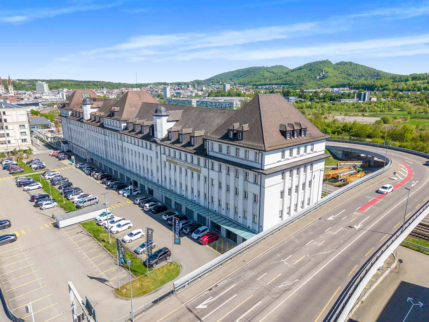 Aerial view of a large white building with a brown roof, surrounded by a parking lot with many cars parked.