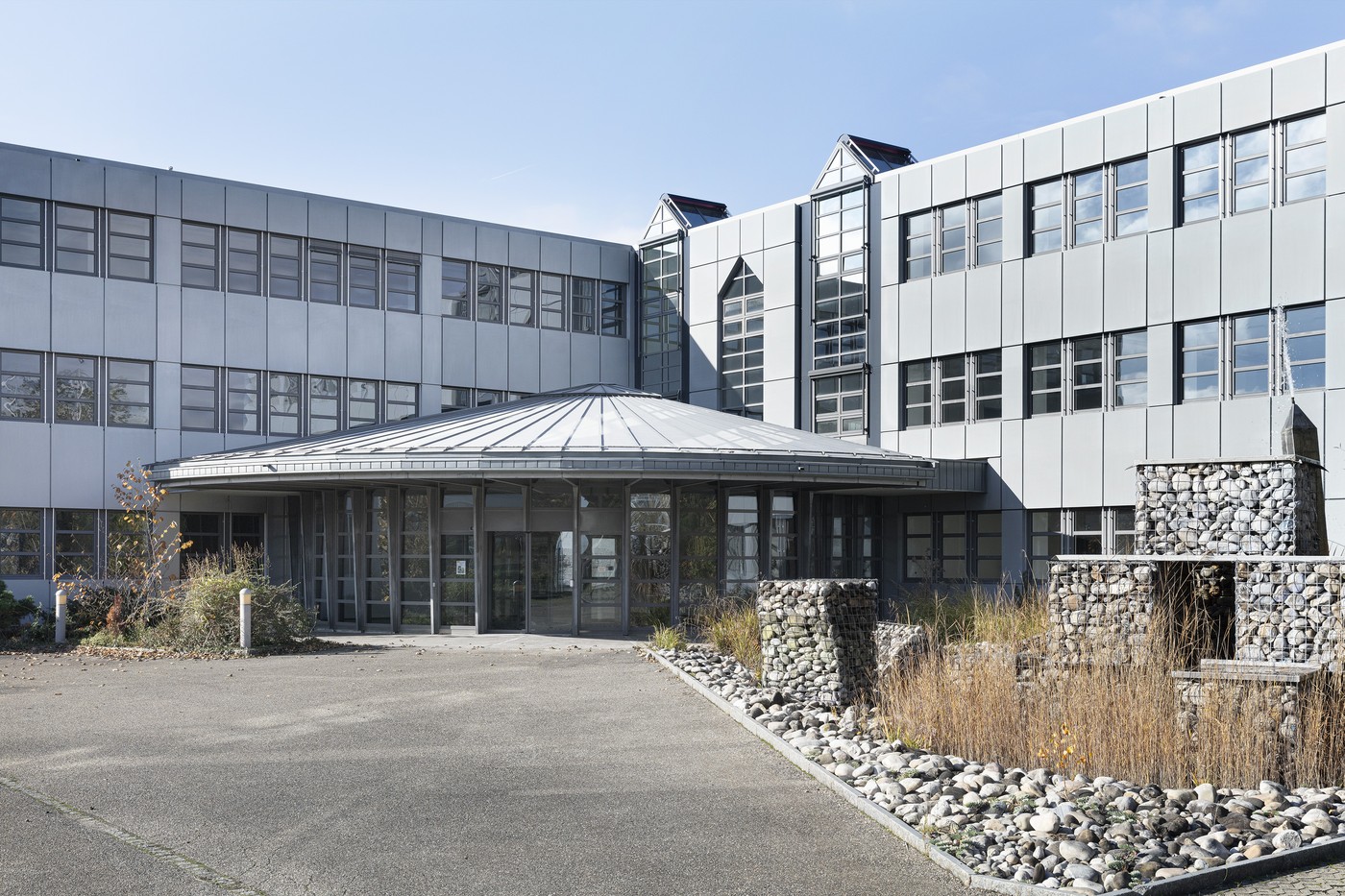 Large modern commercial building with a glass and metal facade, a domed roof structure, and a paved parking area in the foreground. The building has multiple floors and large windows. There is also a gabion wall feature with stones near the entrance.