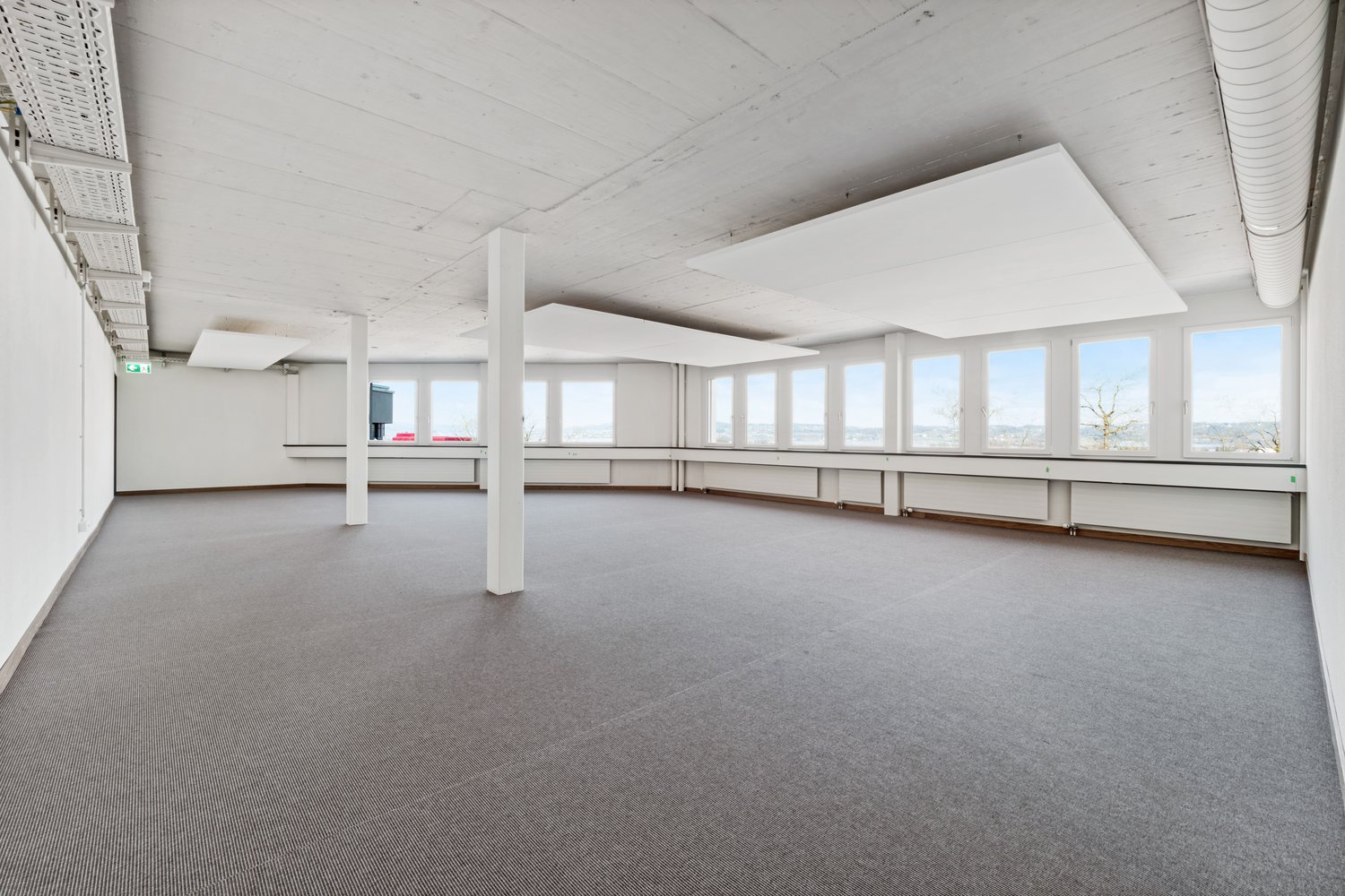 Large open-plan office space with high ceilings, white walls, and large windows providing natural light. The room has a carpeted floor and several support columns. The space appears to be unoccupied and ready for use.