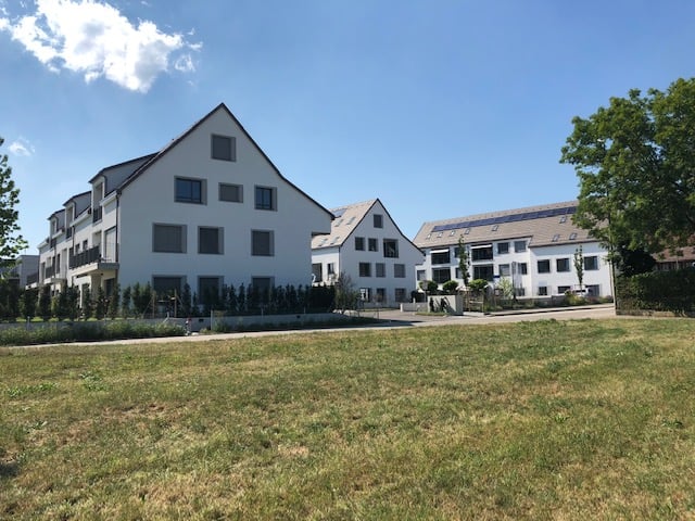Three white apartment buildings with several floors and balconies, large green field in front, solar panels on some roofs, trees and shrubs in the area.
