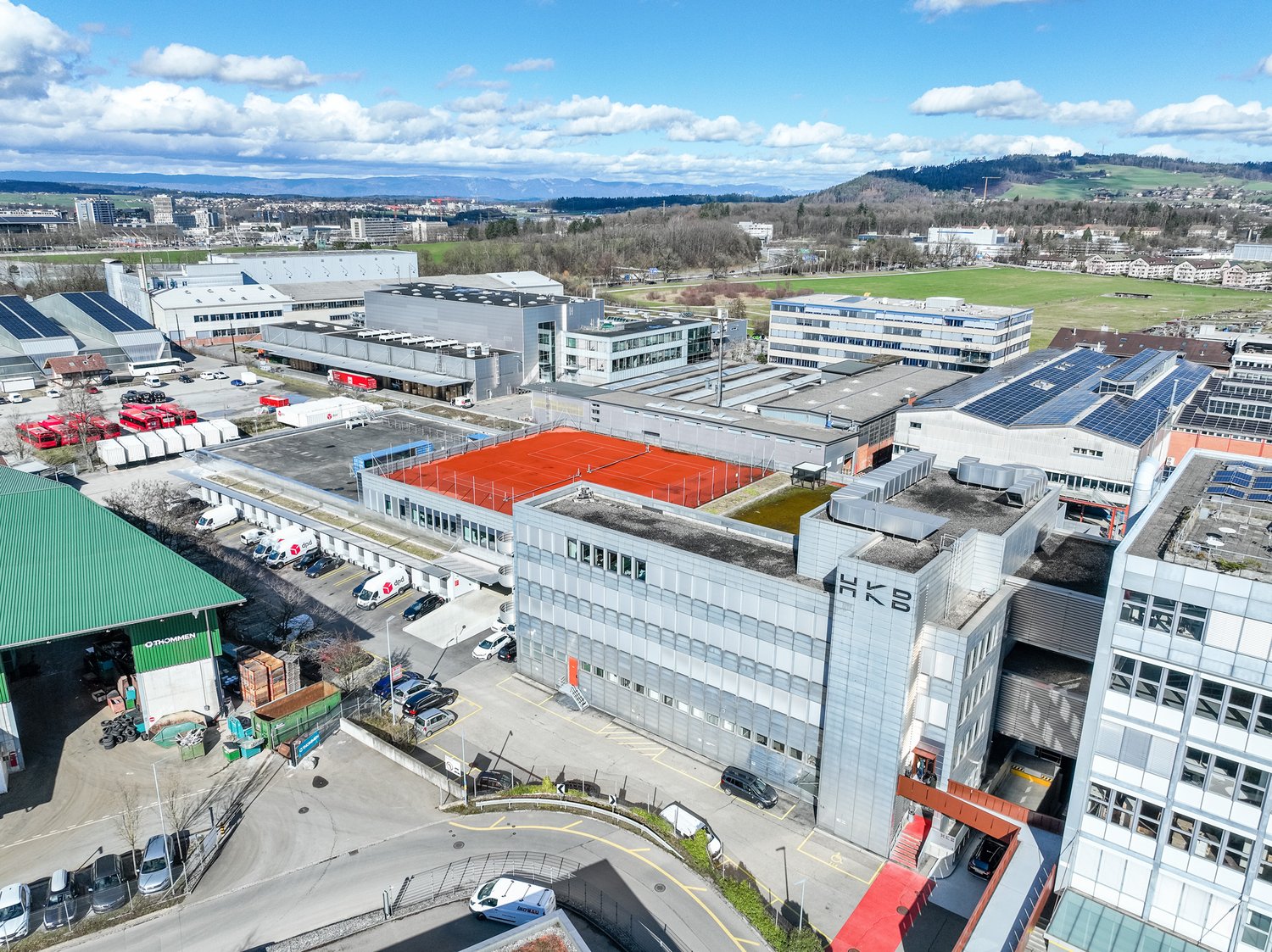 Industrial buildings with parking lots, solar panels on roofs, a red tennis court, multiple buildings, and surrounding area