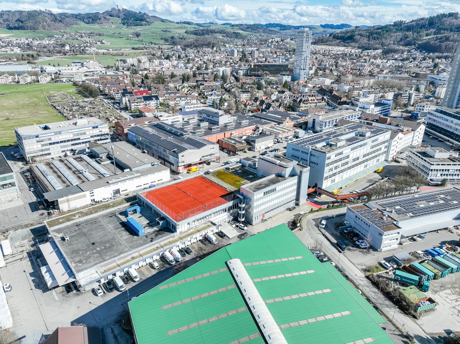 Large industrial area with multiple buildings, some with red tennis courts, multiple solar panels, extensive parking spaces, and multiple cranes and construction vehicles