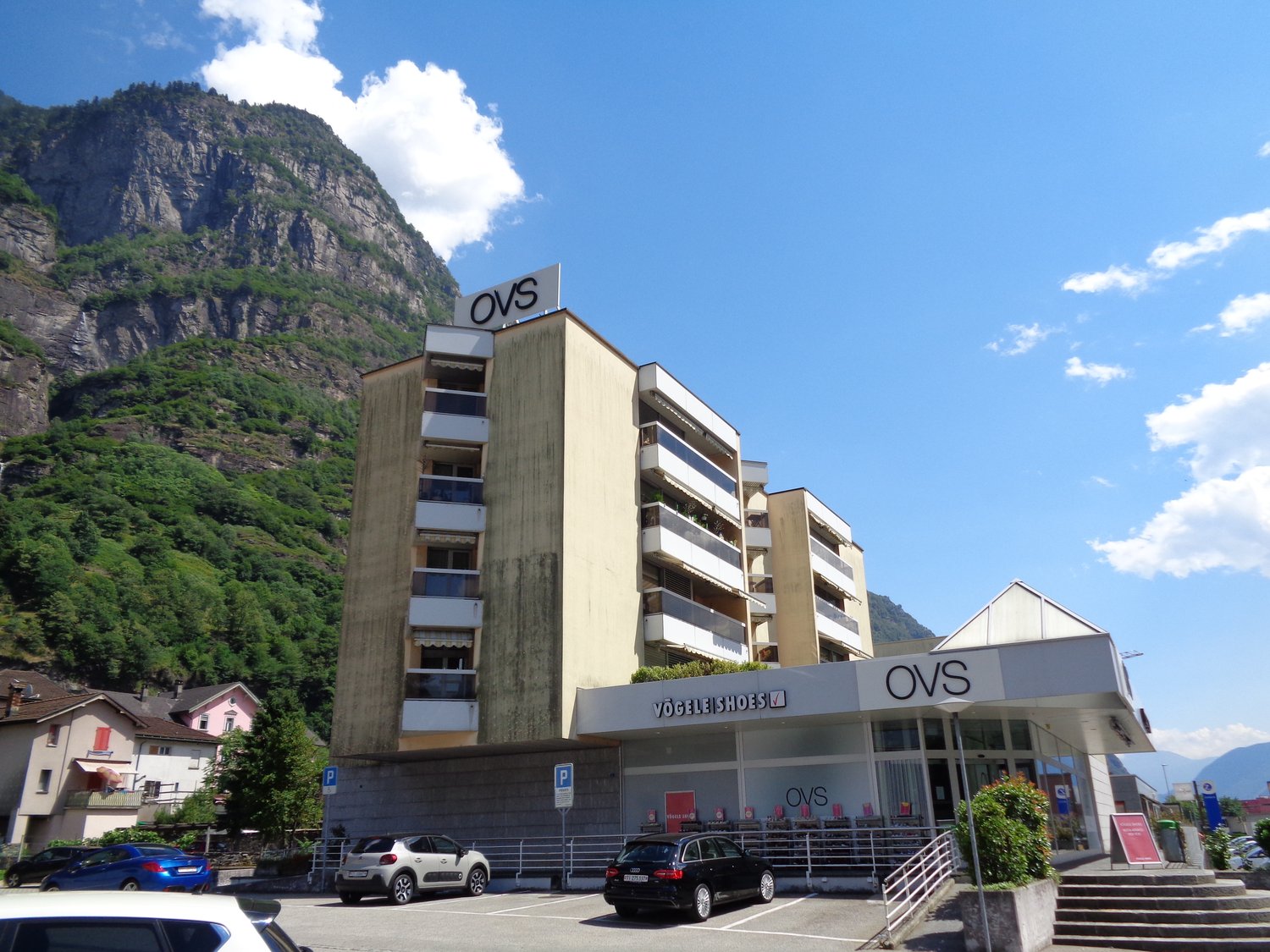 Apartment building with shops on ground floor. Multiple parking spots in front. Mountain backdrop.