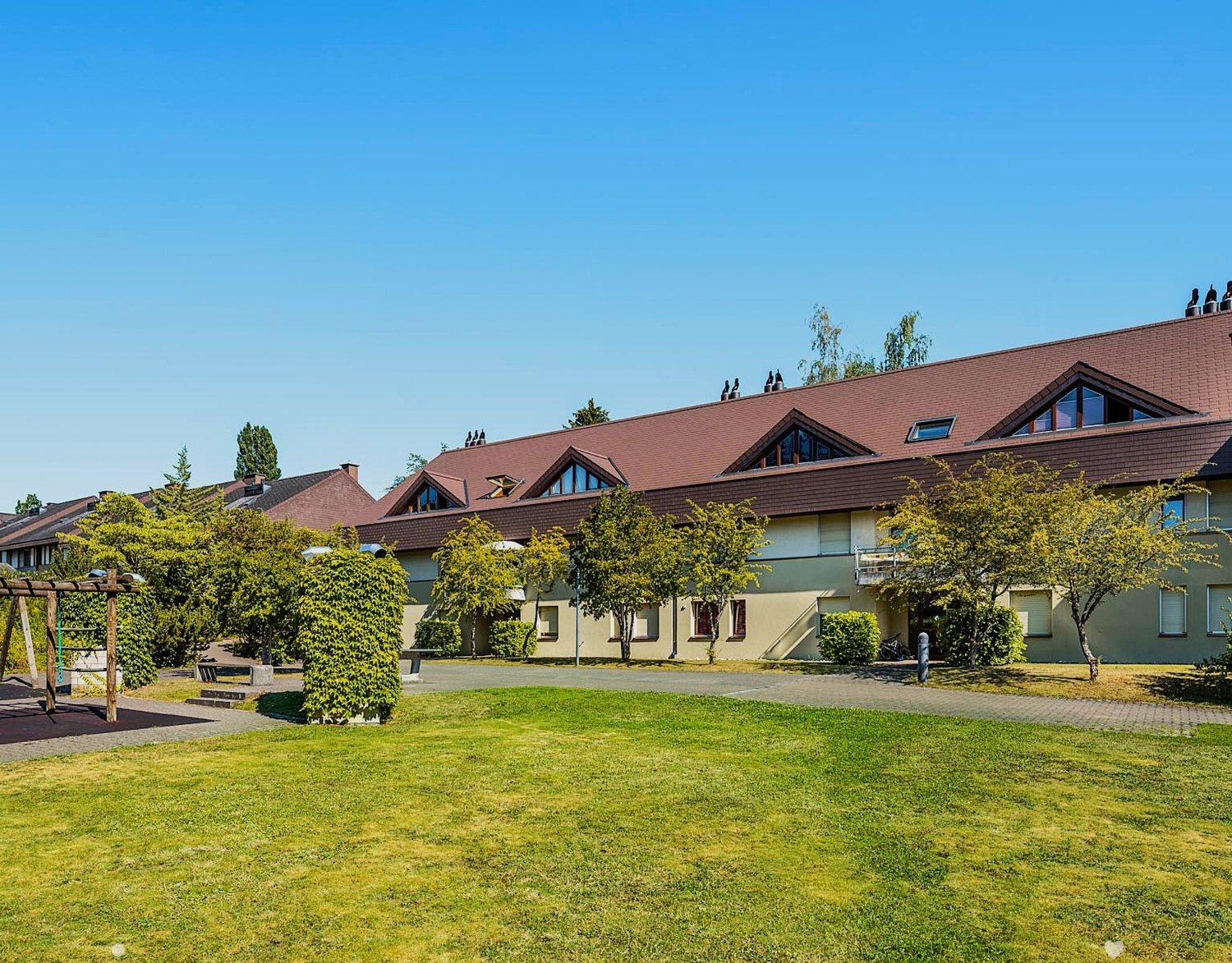 Multiple story building with many windows, brown roof, wide front yard with greenery and playground