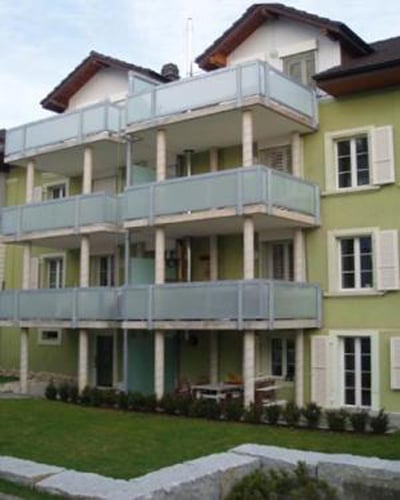 A modern, green-painted building with several floors and multiple balconies. The balconies are covered with glass railings. The building has several windows with white shutters. A well-kept lawn with bushes and a walkway can be seen in front of the buildi