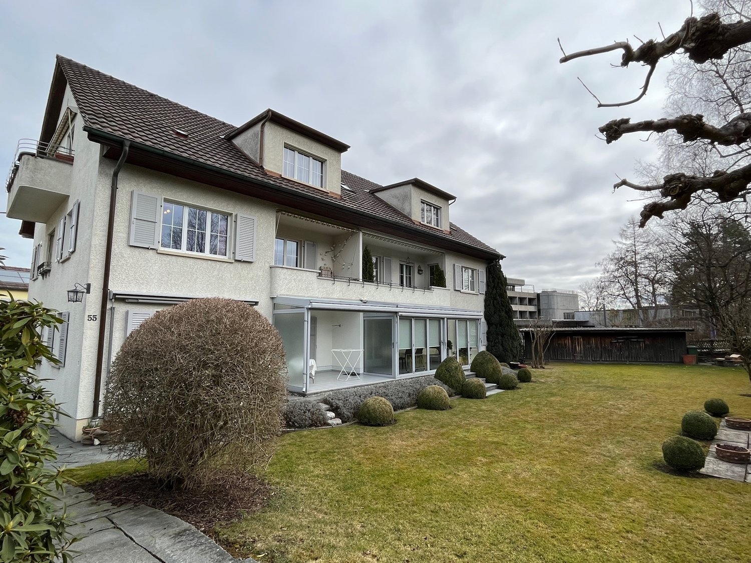 Two story house, brown roof, glass walls, balcony, lawn, garden, trees
