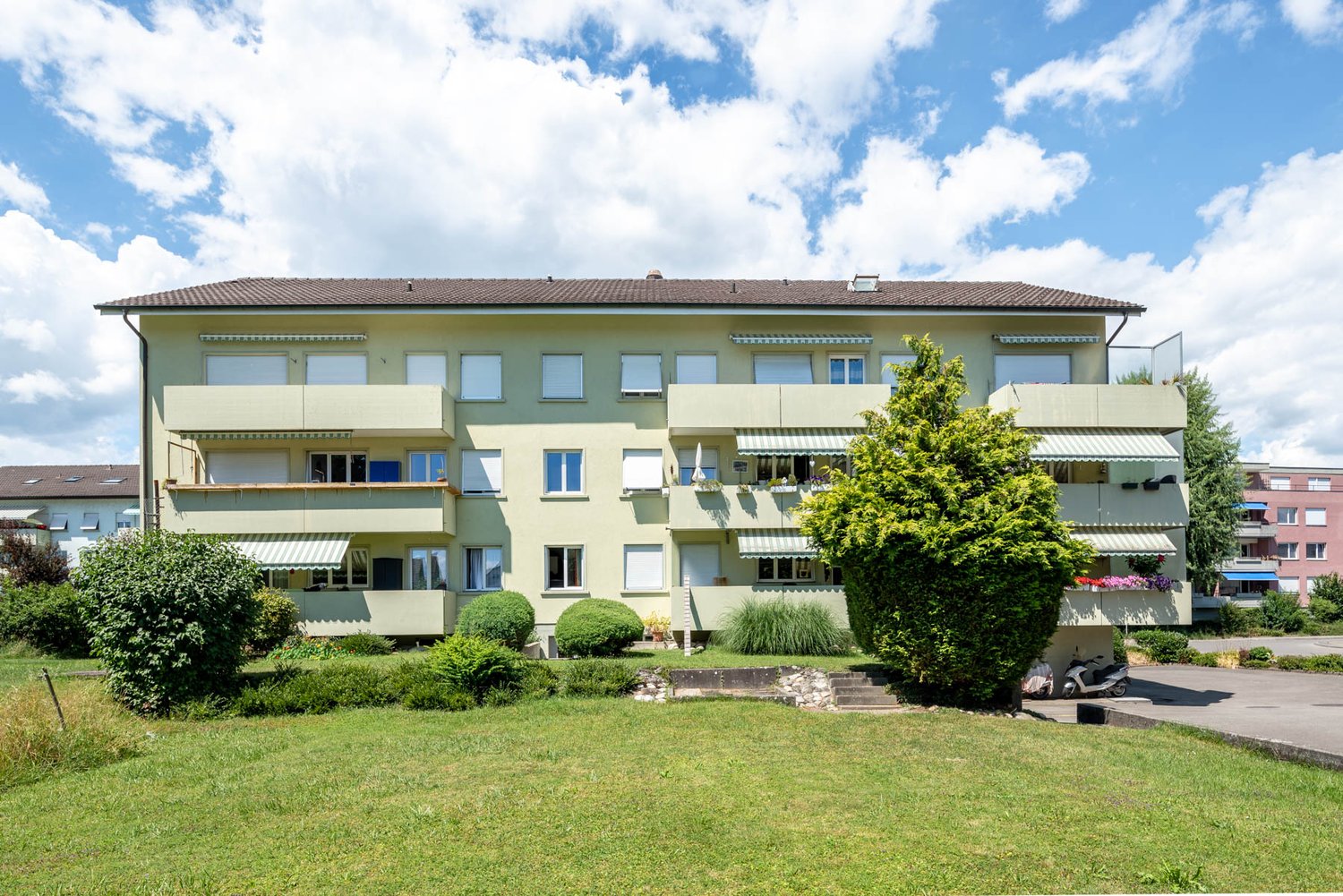 3 story apartment building, beige exterior, brown roof, multiple windows, awnings, balconies, front yard, pathway