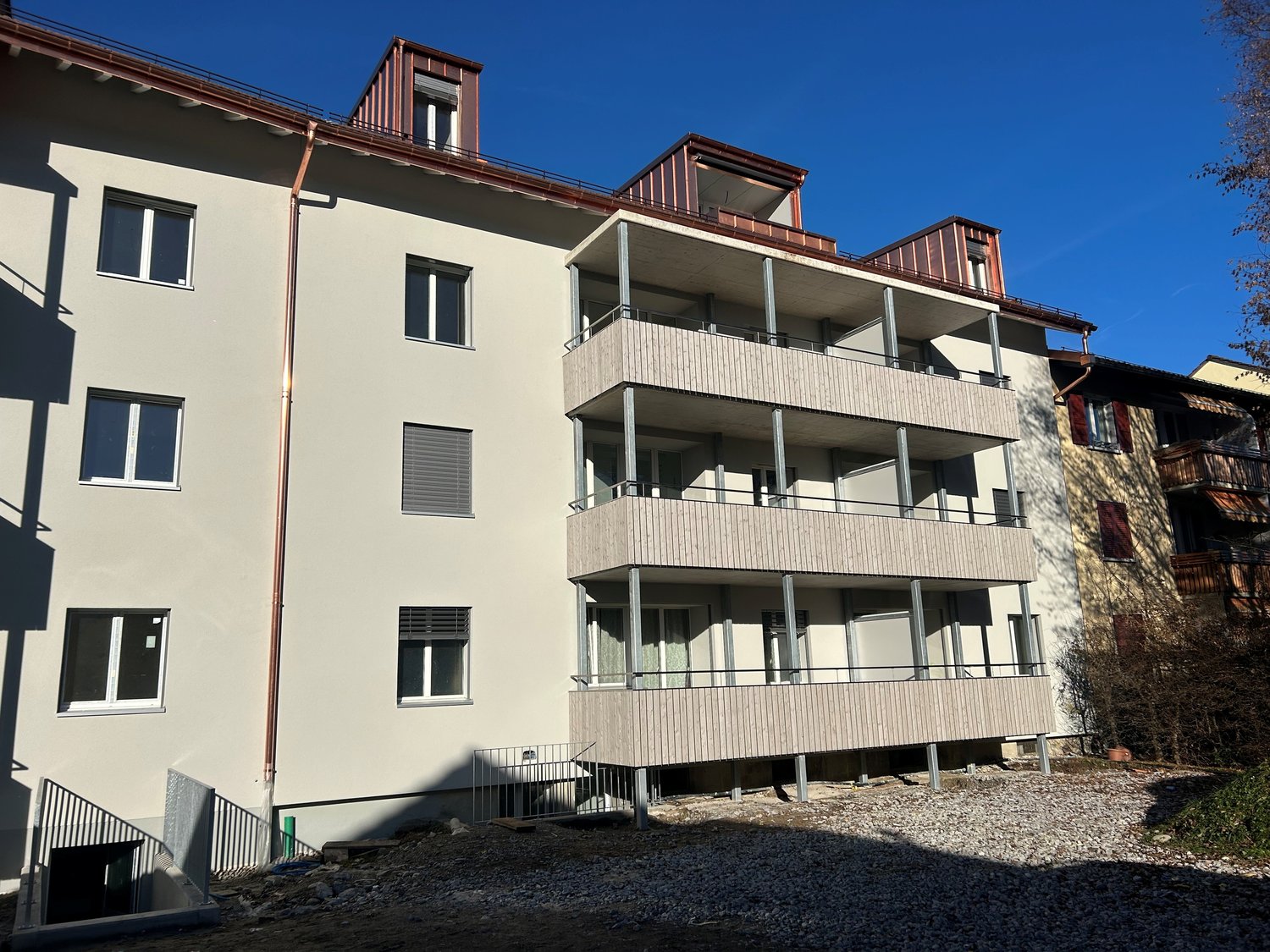 Modern apartment building, white exterior, balconies on upper floors, multiple windows, metal railings, concrete ground floor