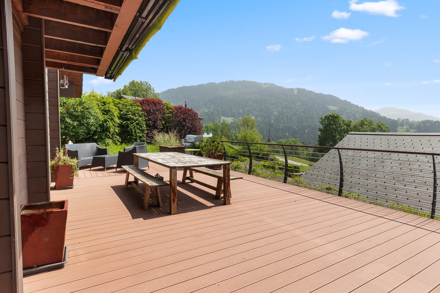 Wooden deck with a dining table and chairs, surrounded by lush greenery and overlooking a scenic mountain landscape with a blue sky and clouds