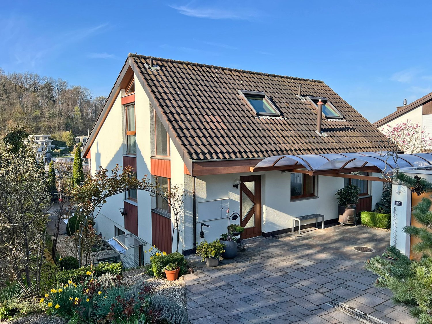 Detached house, brown roof, white walls, brown trim, two windows, two dormers, covered entrance, paved driveway, garden with plants and flowers