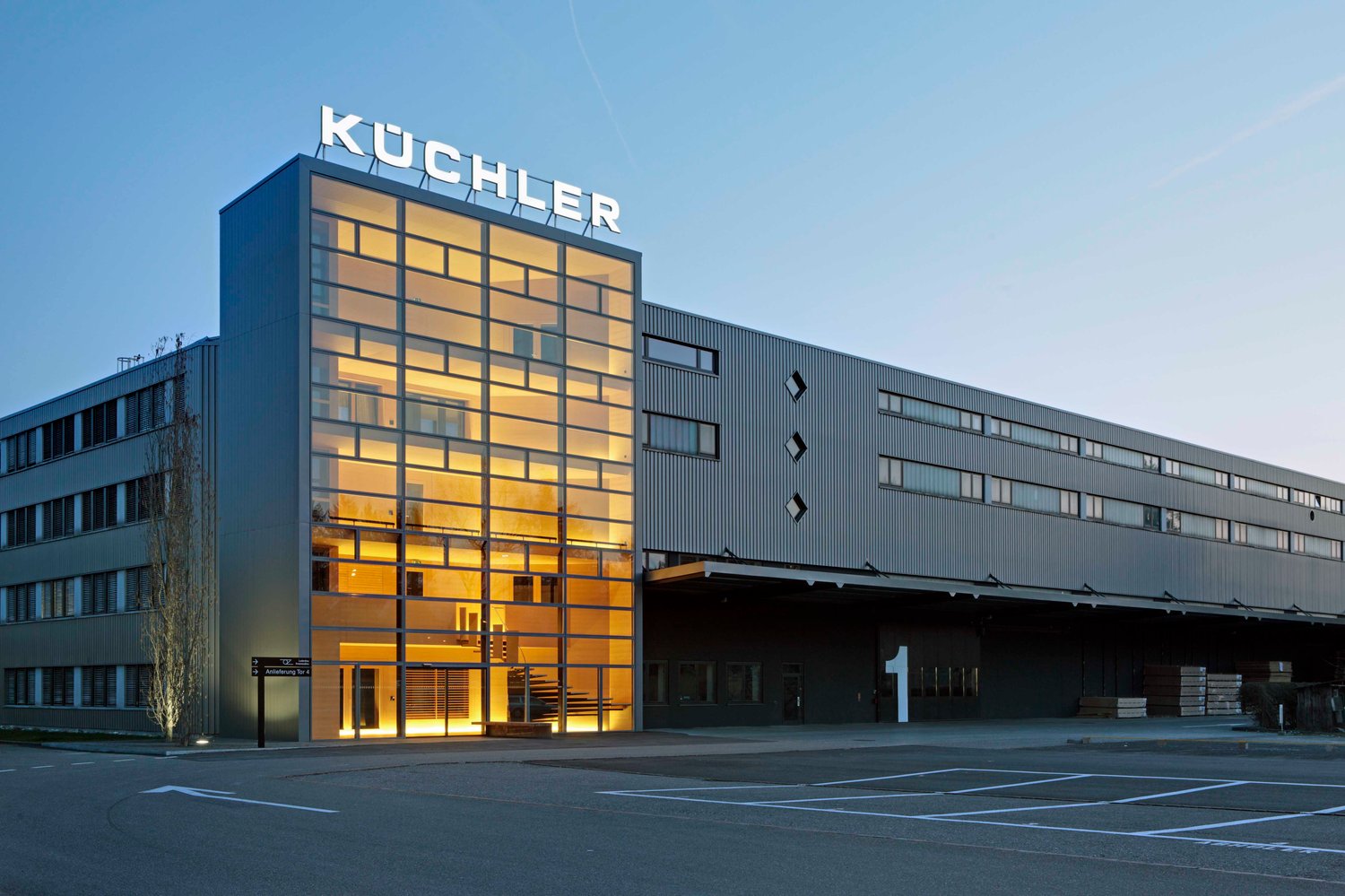 Large modern building, glass walls, metal panels, ample parking spaces, entrance with staircase