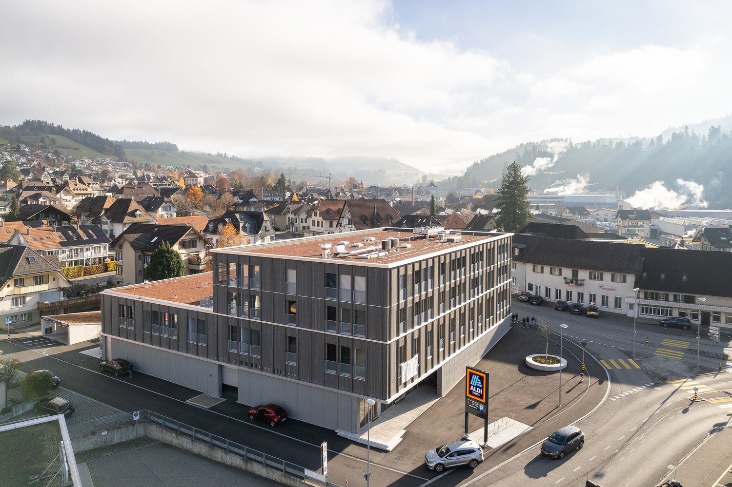 A modern multi-story office building with a gray exterior and large windows, situated in a town with traditional architecture and surrounded by hills and forests.
